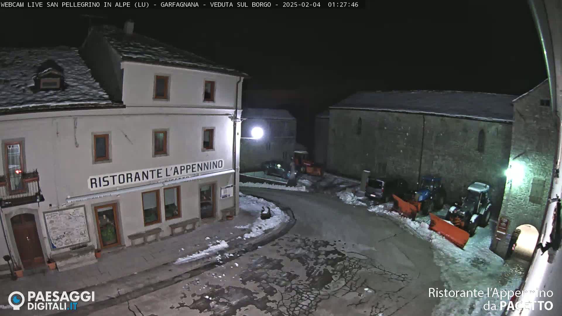 A snow-covered town square at night, illuminated by streetlights, features a restaurant and several parked vehicles and snowplows.