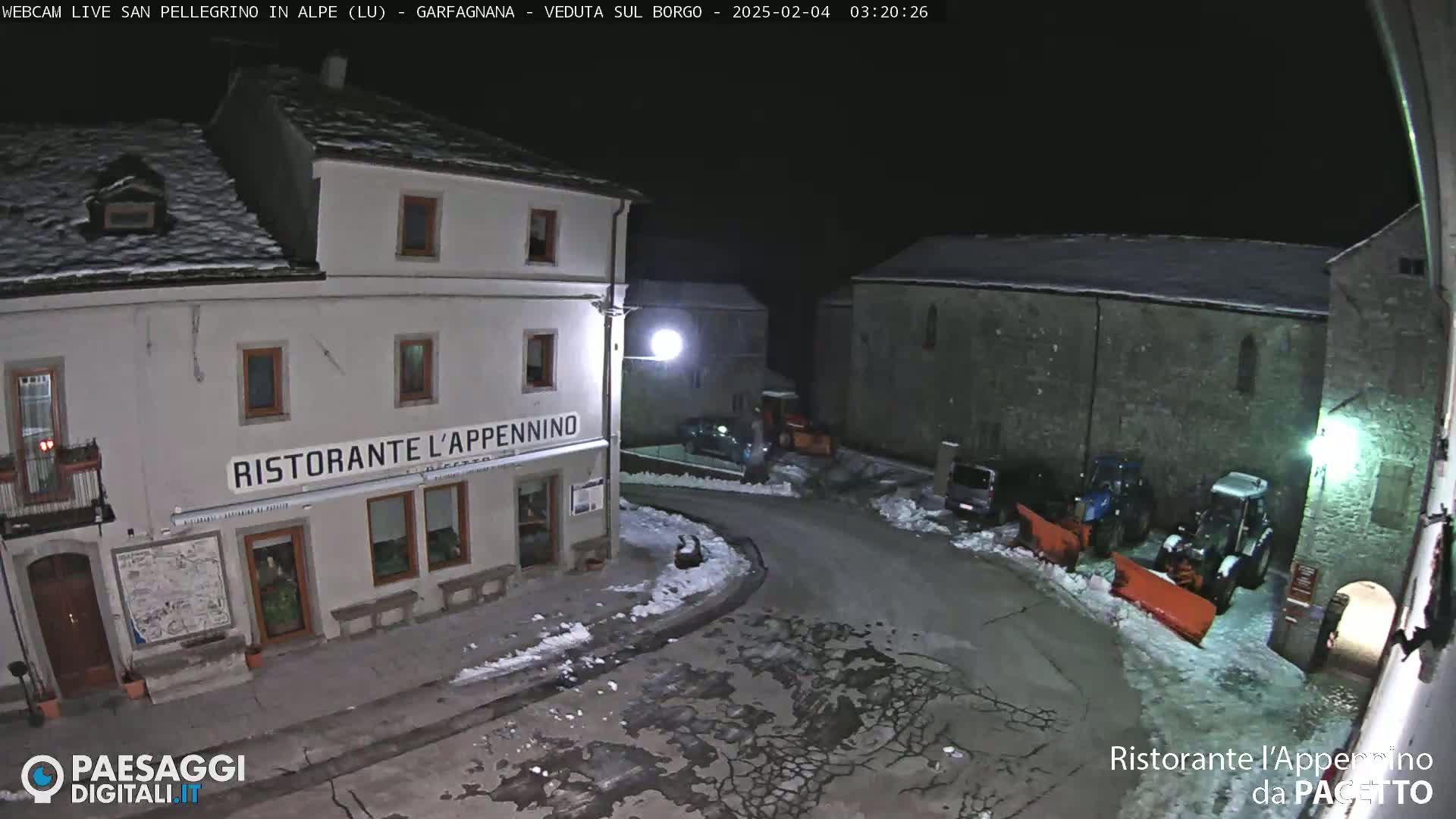 A snow-covered village square at night, illuminated by streetlights, features a restaurant and parked vehicles, with snow plows present.