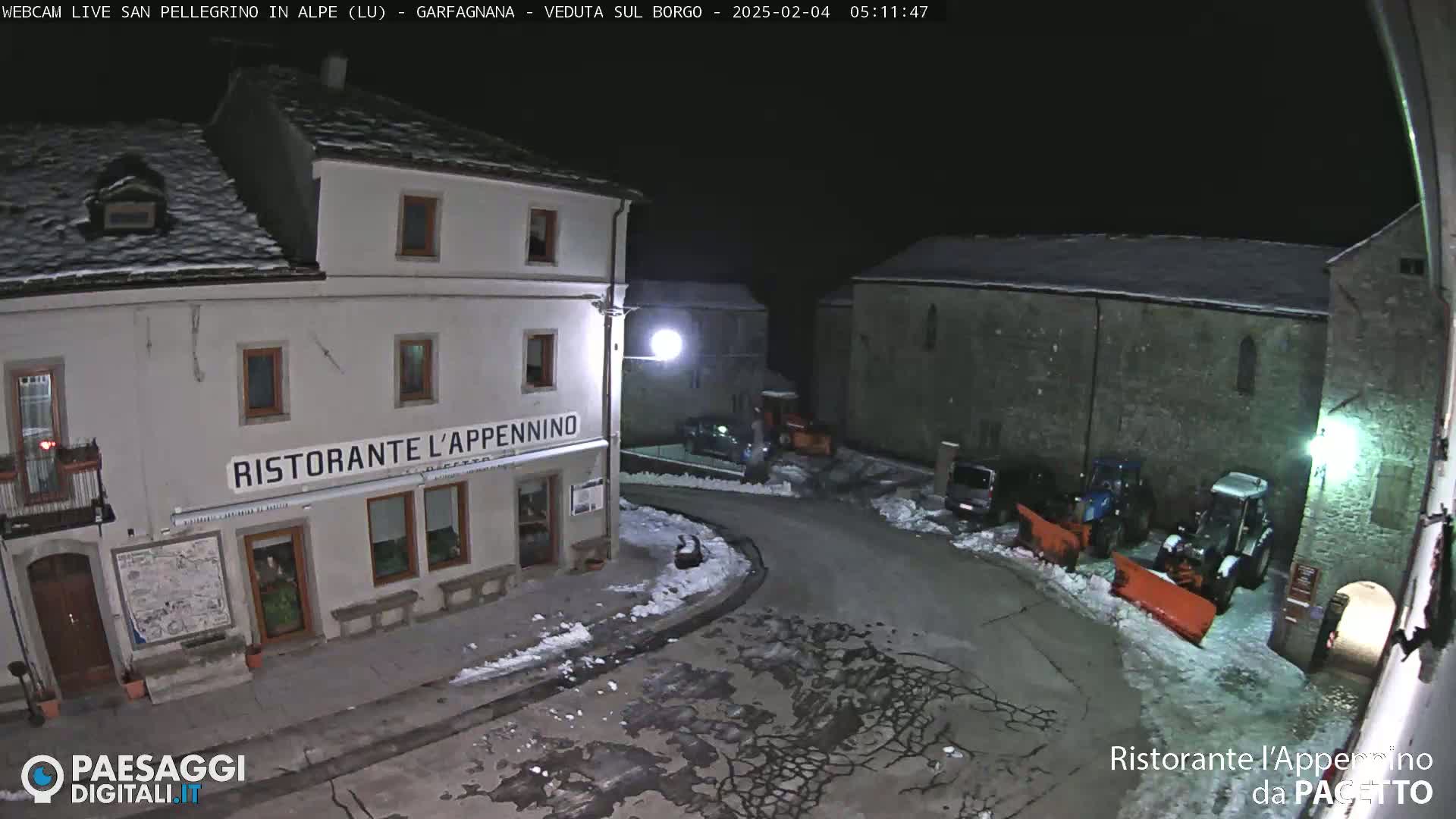 A snow-covered town square at night, illuminated by streetlights, shows several buildings, parked cars, and snowplows.