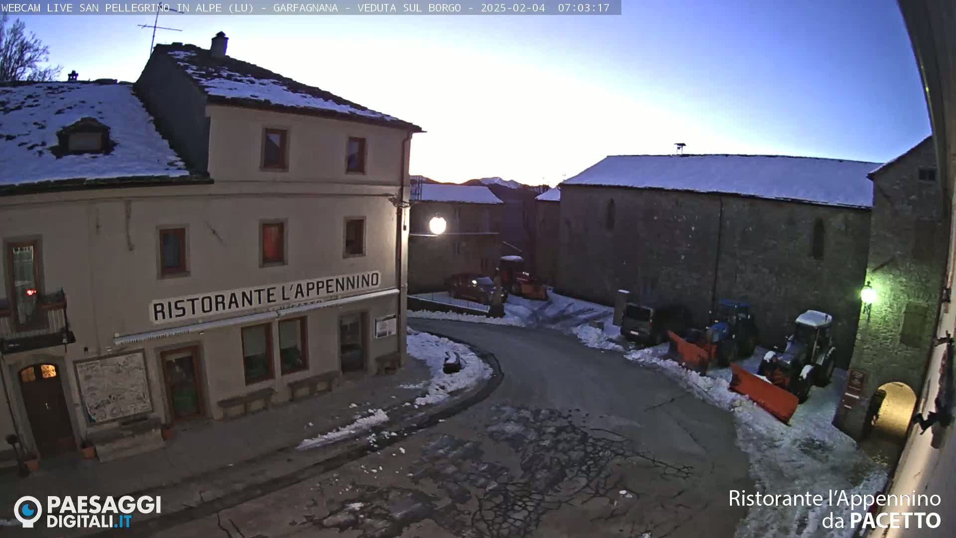 A snow-covered village square at twilight, with several buildings, some parked vehicles, and snow plows.