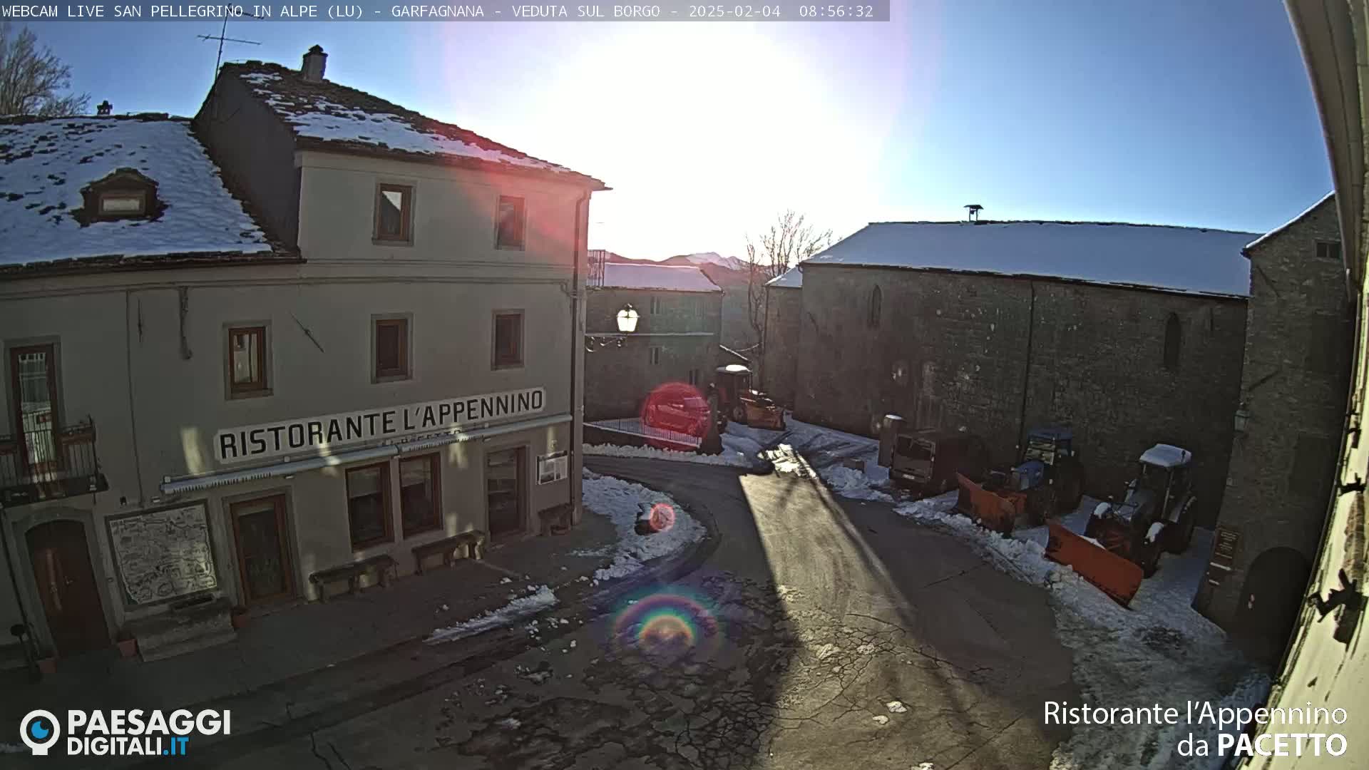 A sunny, snowy village square with a restaurant, several stone buildings, and snowplows parked nearby.