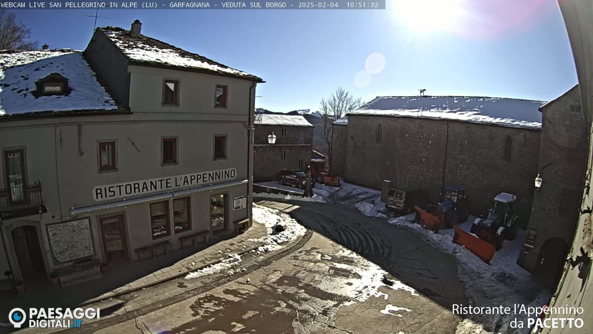 A sunny, snowy village square with a restaurant and several snow-clearing vehicles parked nearby.