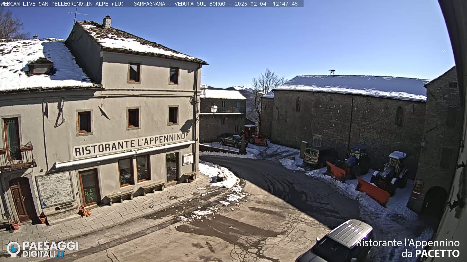 A sunny, snow-dusted village square with several buildings, some vehicles, and snow-clearing equipment.