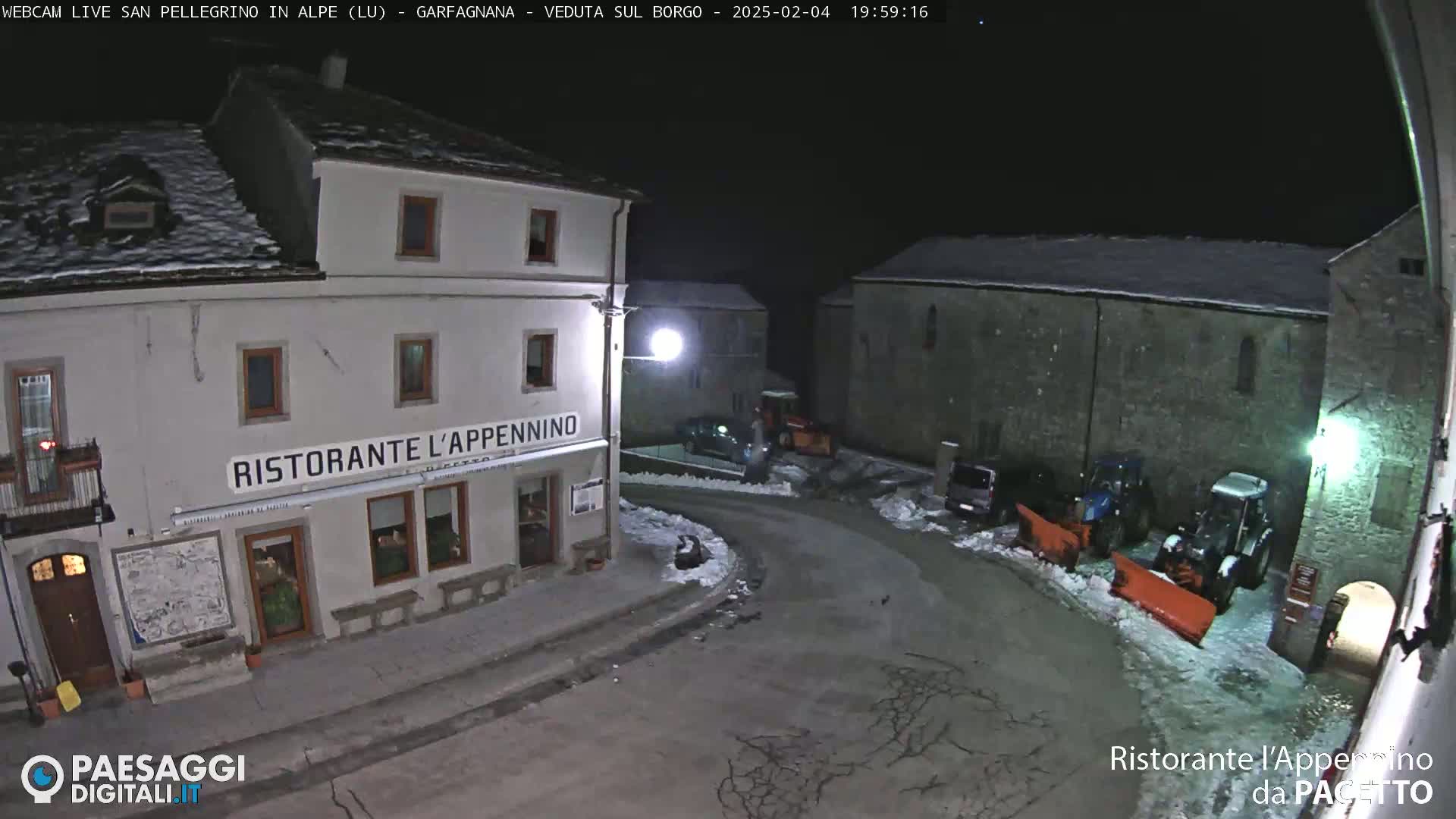 A nighttime, snow-covered village square scene with a restaurant, several parked vehicles, and snowplows.