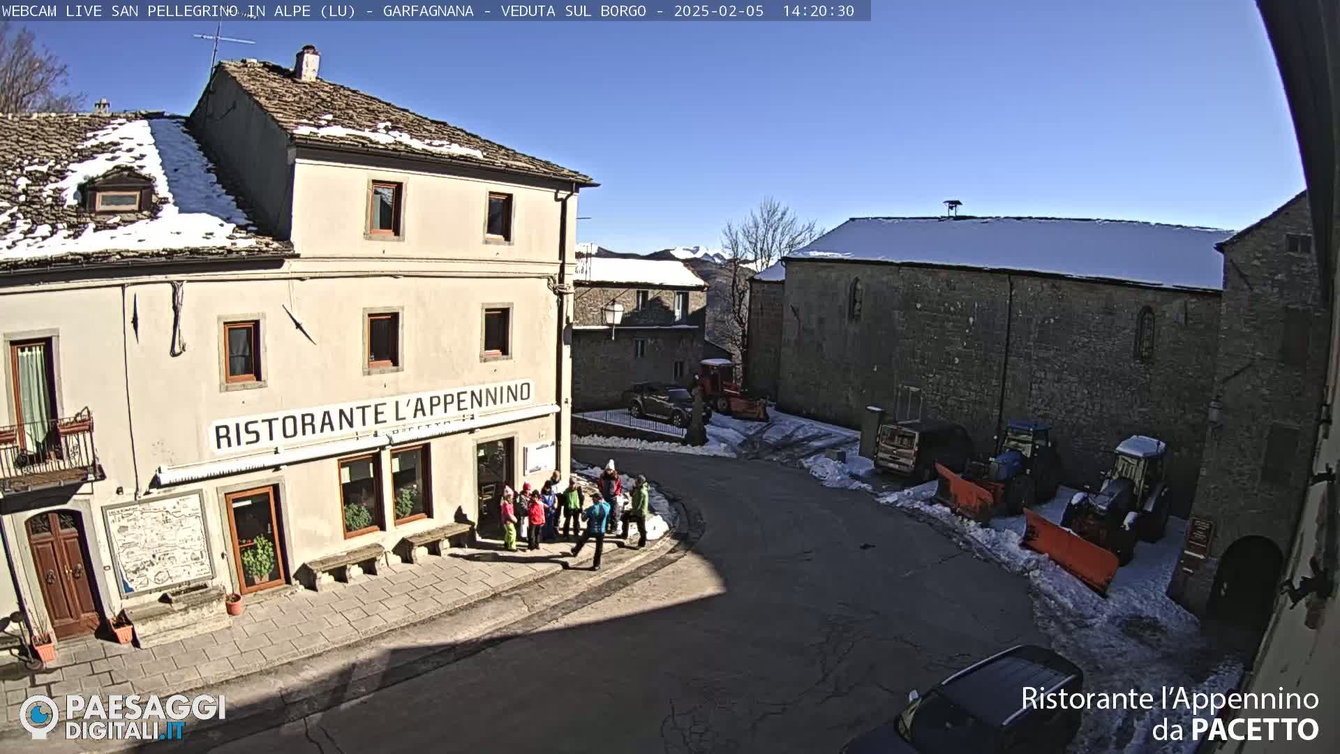 A sunny, snow-dusted village square features a restaurant, several parked vehicles including snowplows, and a group of people gathered near a building.