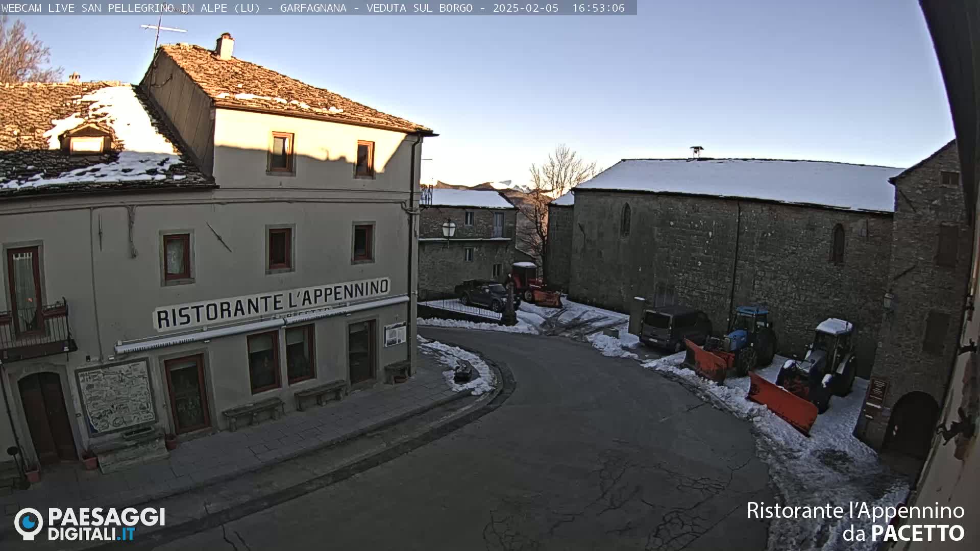 A snow-covered village street scene shows several buildings, some parked vehicles, and snow removal equipment under a clear, bright sky.