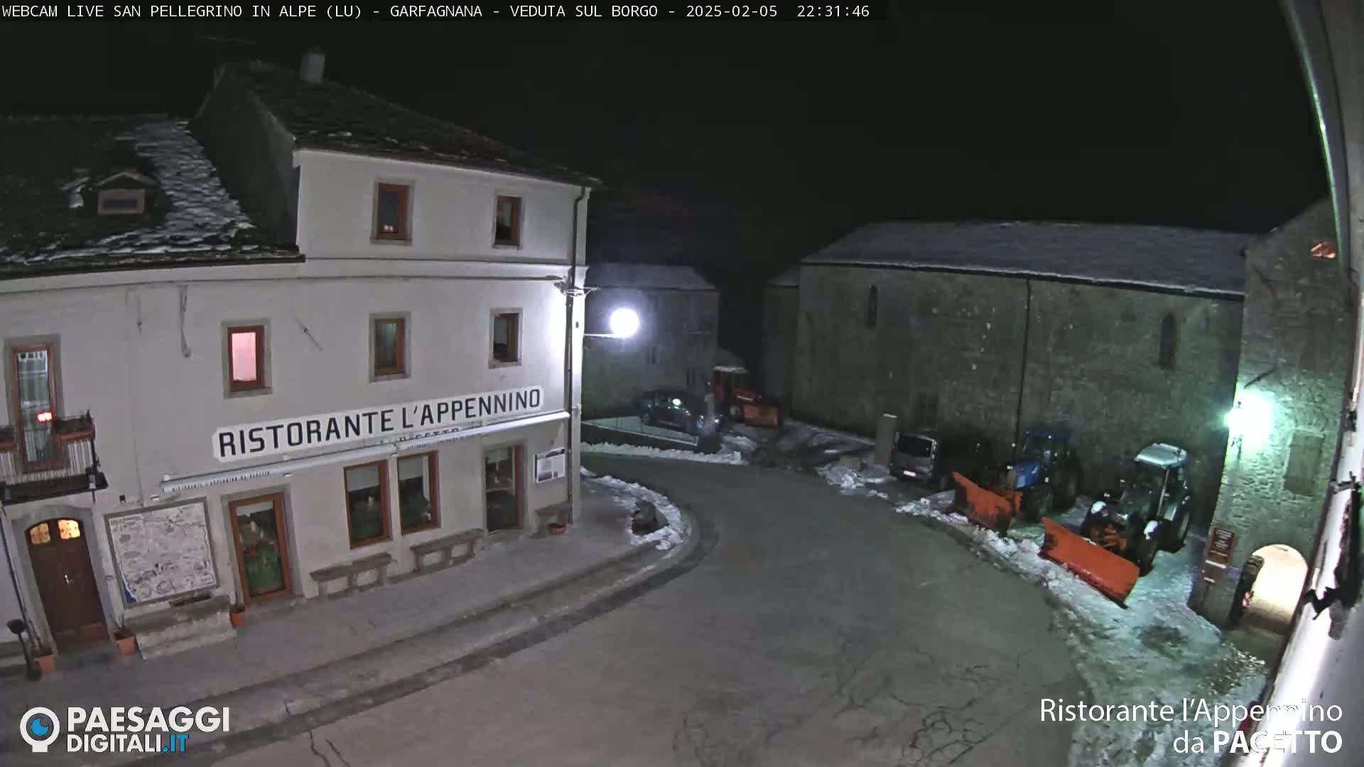 A nighttime view of a snow-dusted village square with a restaurant, several parked vehicles, and snowplows.
