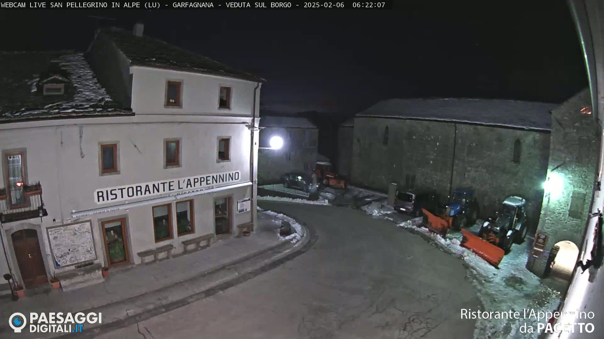 A snow-covered town square at night, illuminated by streetlights, features a restaurant and several vehicles, including snowplows.