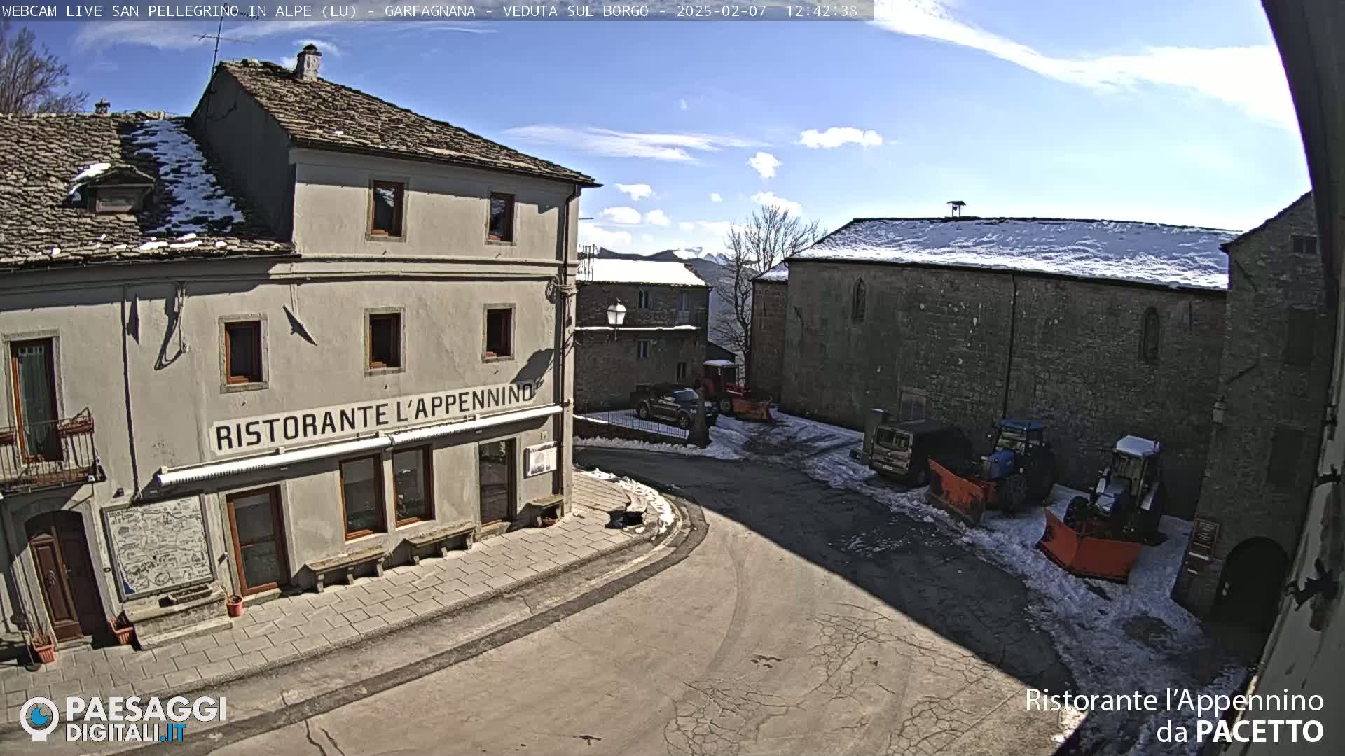 A sunny day with patches of snow on the ground shows a village square with several buildings, including a restaurant, and some snow removal equipment parked nearby.