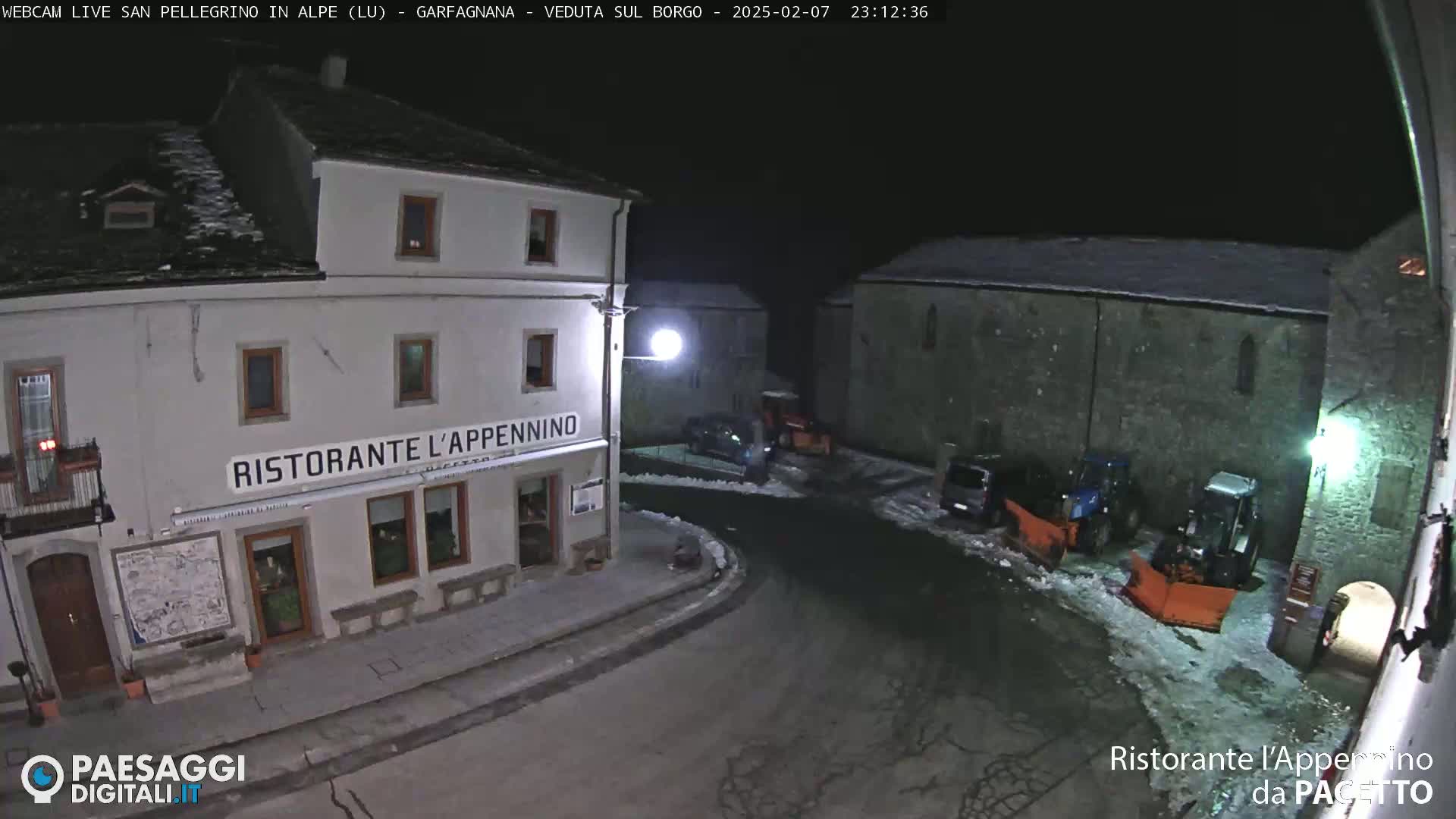 A snow-covered town square at night, illuminated by streetlights, shows a restaurant and several vehicles including snowplows.