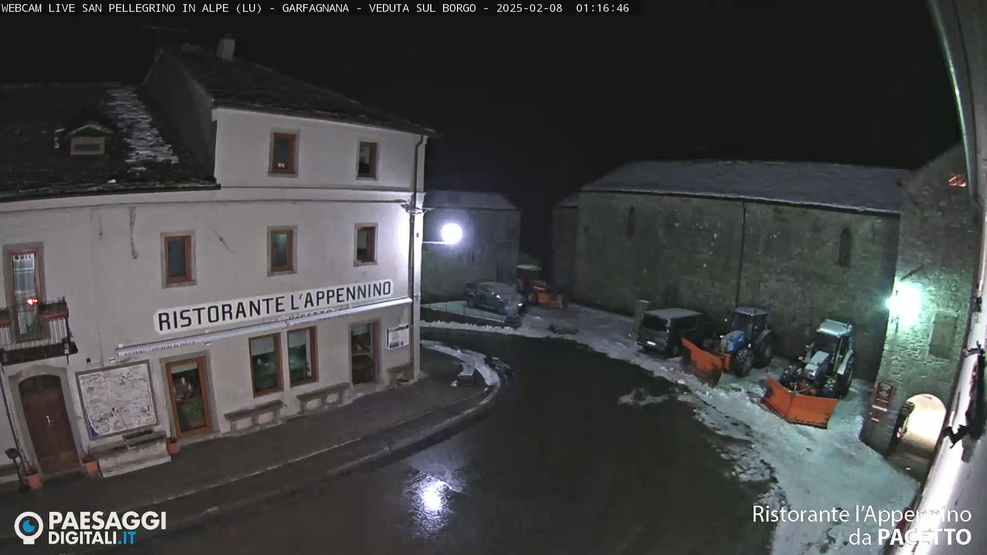A nighttime view of a snow-covered town square with several buildings, including a restaurant, and some snow removal equipment.
