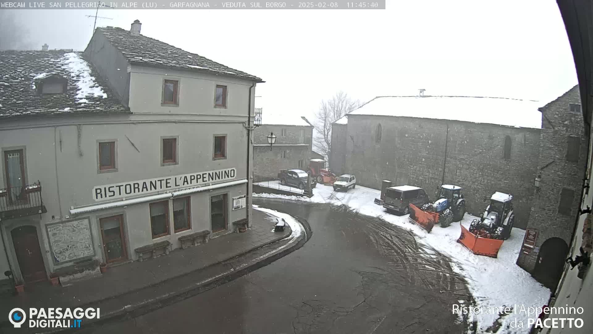 A snow-covered village square with several vehicles, including snow plows, parked near a restaurant.