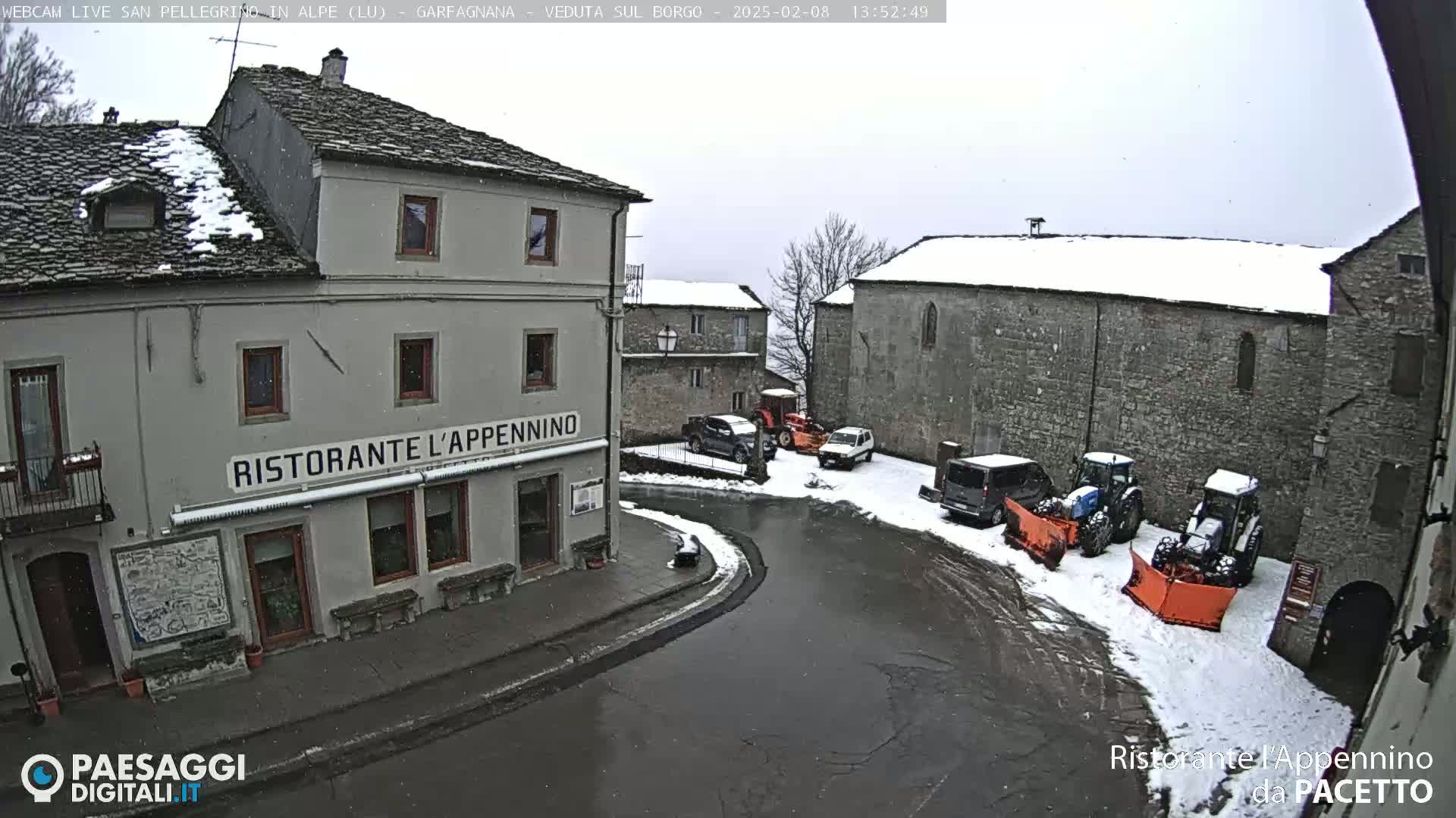 A snow-covered village square with several vehicles, including tractors and snowplows, parked near stone buildings under a light snowfall.
