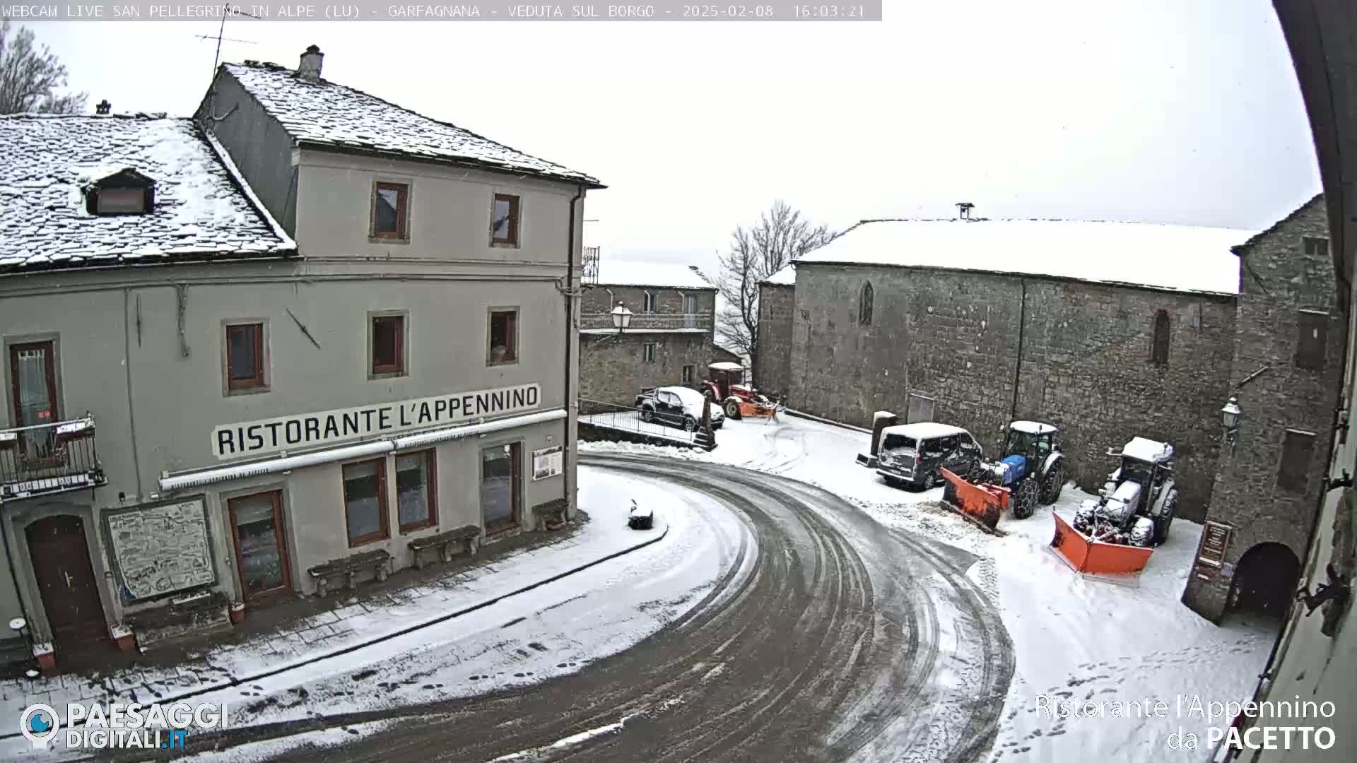 A snow-covered village square with several buildings, parked vehicles, and snow-clearing equipment.