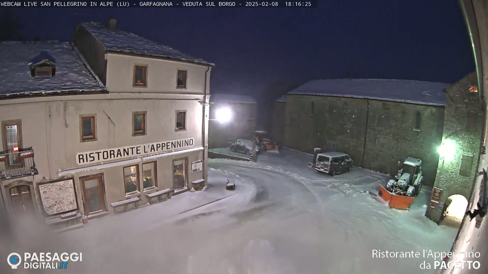 A snow-covered town square at night, with a restaurant, parked vehicles, and snow-clearing equipment.