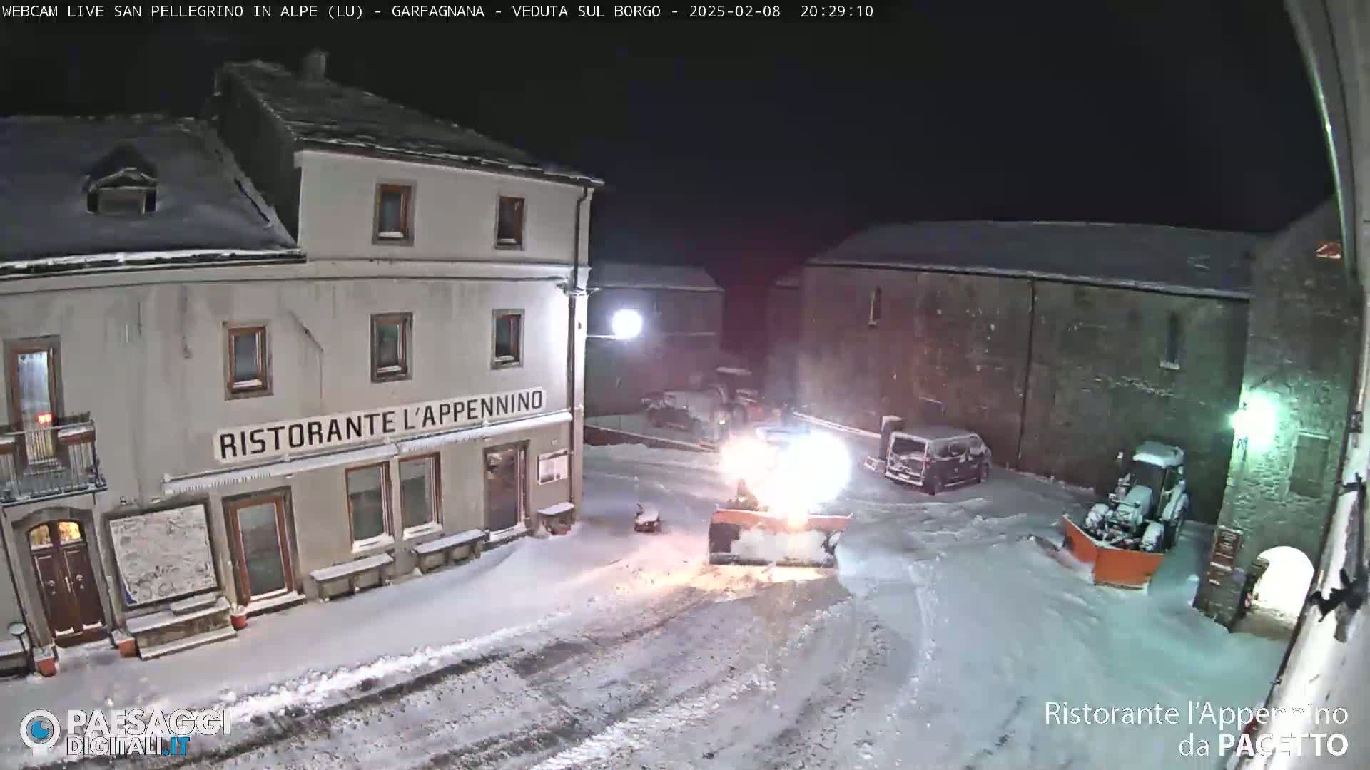 A snowplow clears snow from a town square at night, near buildings including a restaurant.