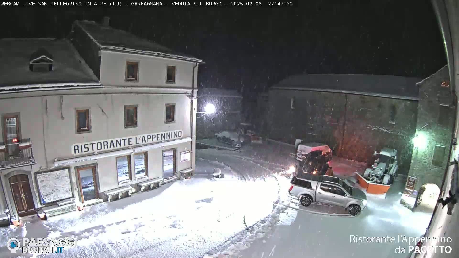 A snow-covered town square at night, with a restaurant, several vehicles, and a snow plow in view amidst falling snow.