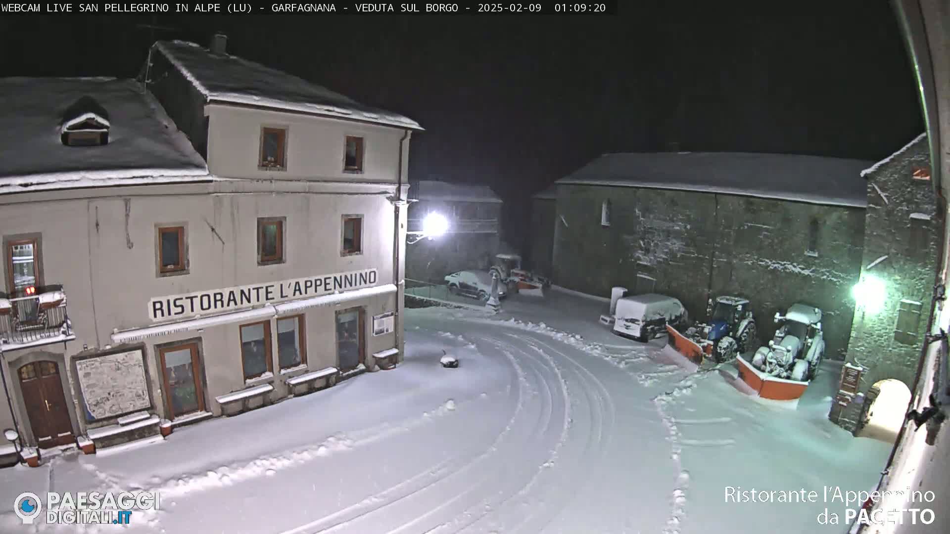 A snow-covered town square at night, illuminated by streetlights, shows a restaurant and snowplows parked near stone buildings.