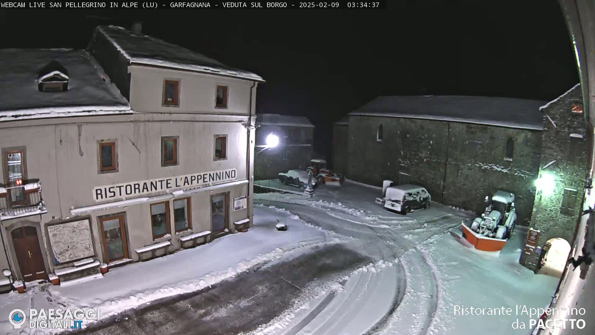 A snow-covered town square at night, with a restaurant, several buildings, and snow-clearing equipment.