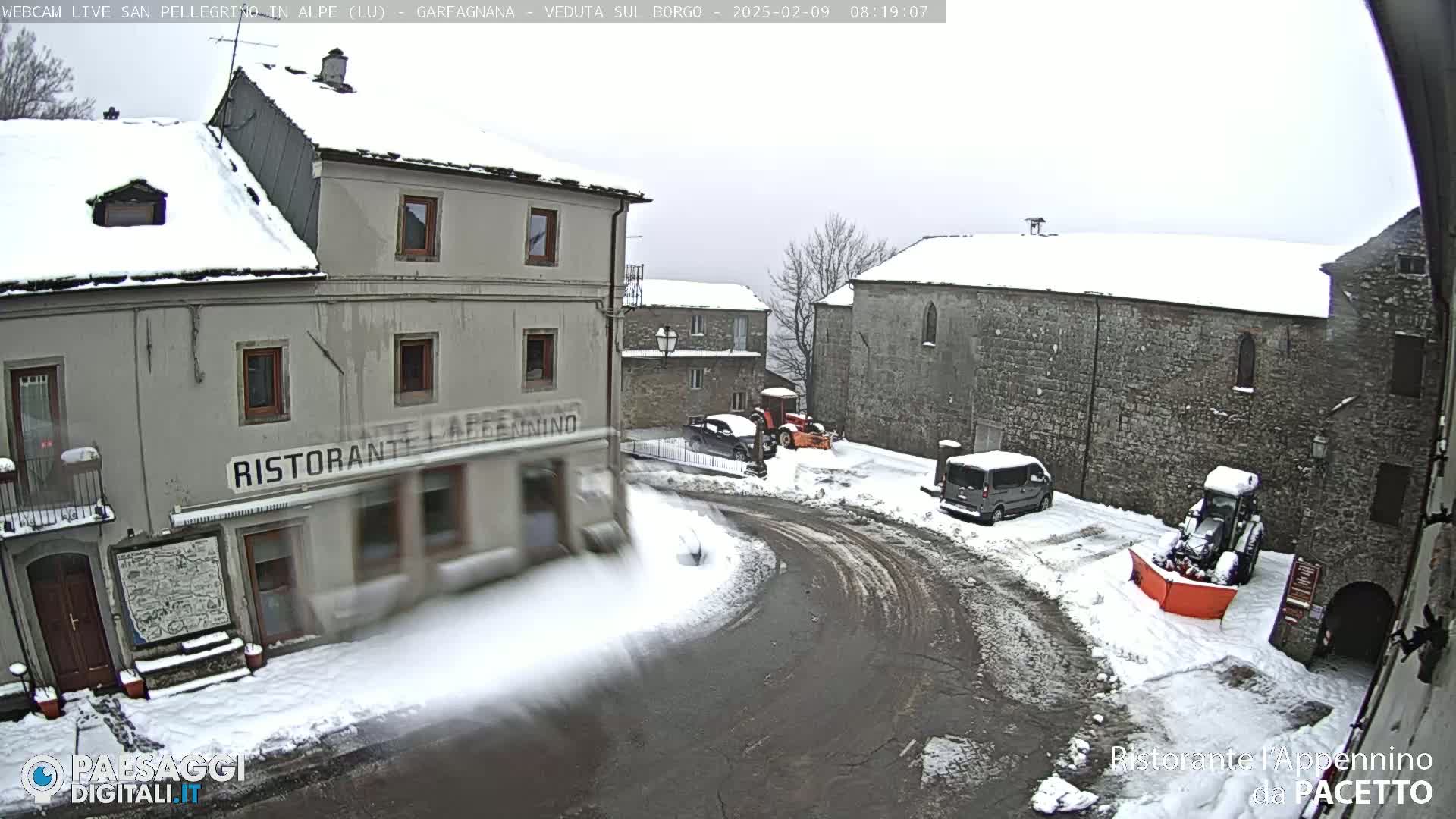 A snow-covered village square with several buildings, a restaurant, parked vehicles, and snow-clearing equipment.