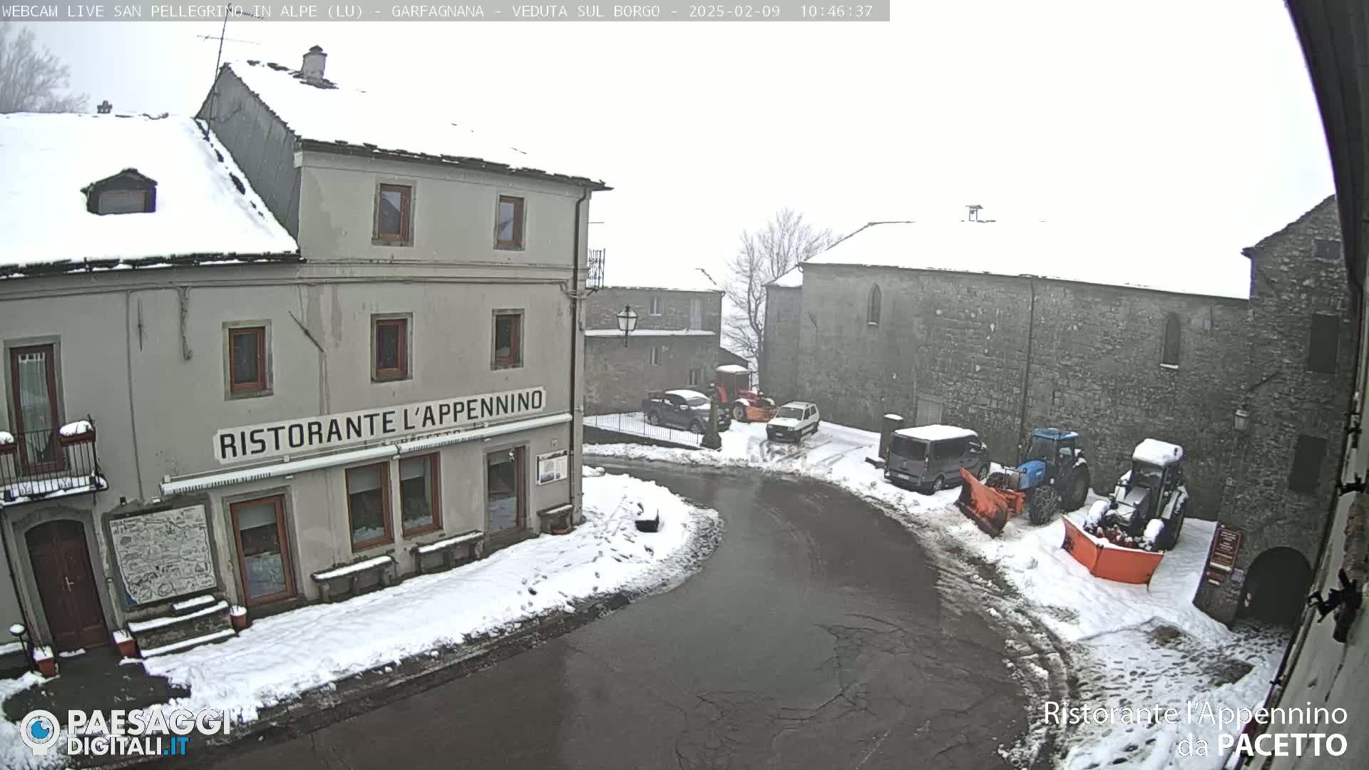 A snow-covered village square with several vehicles, including snow plows, parked near stone buildings, one of which is a restaurant.