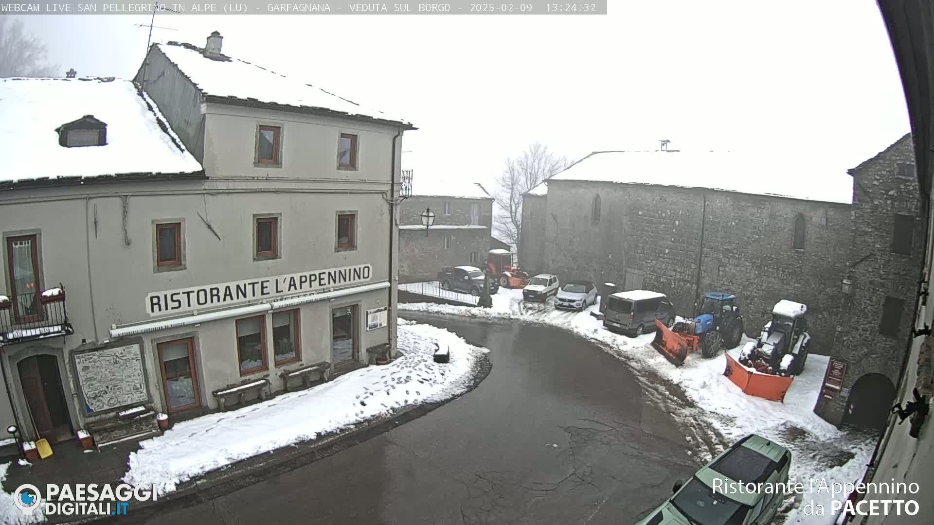 A snow-covered village square with several cars and snowplows parked around a restaurant.