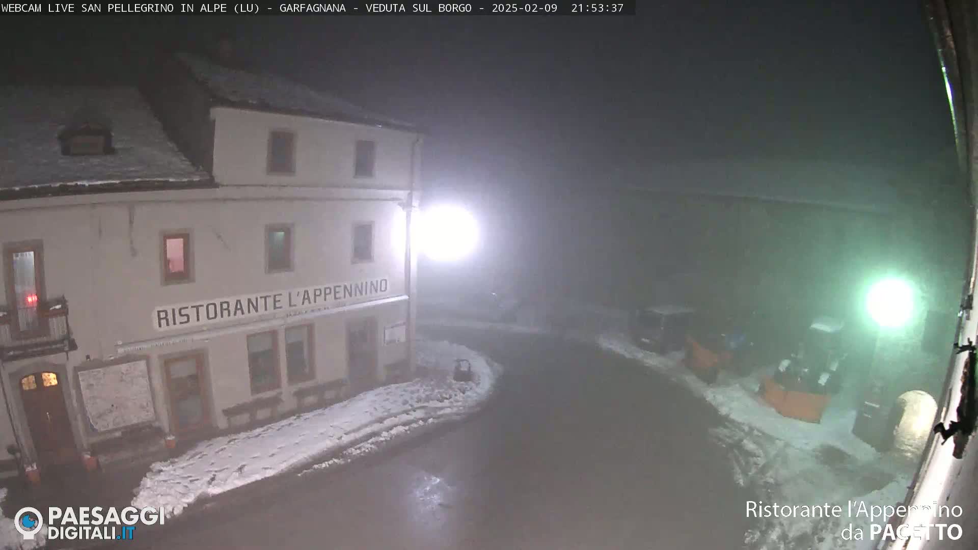 A nighttime, snowy scene shows a restaurant called Ristorante L'Appennino, with some vehicles parked nearby under hazy conditions.