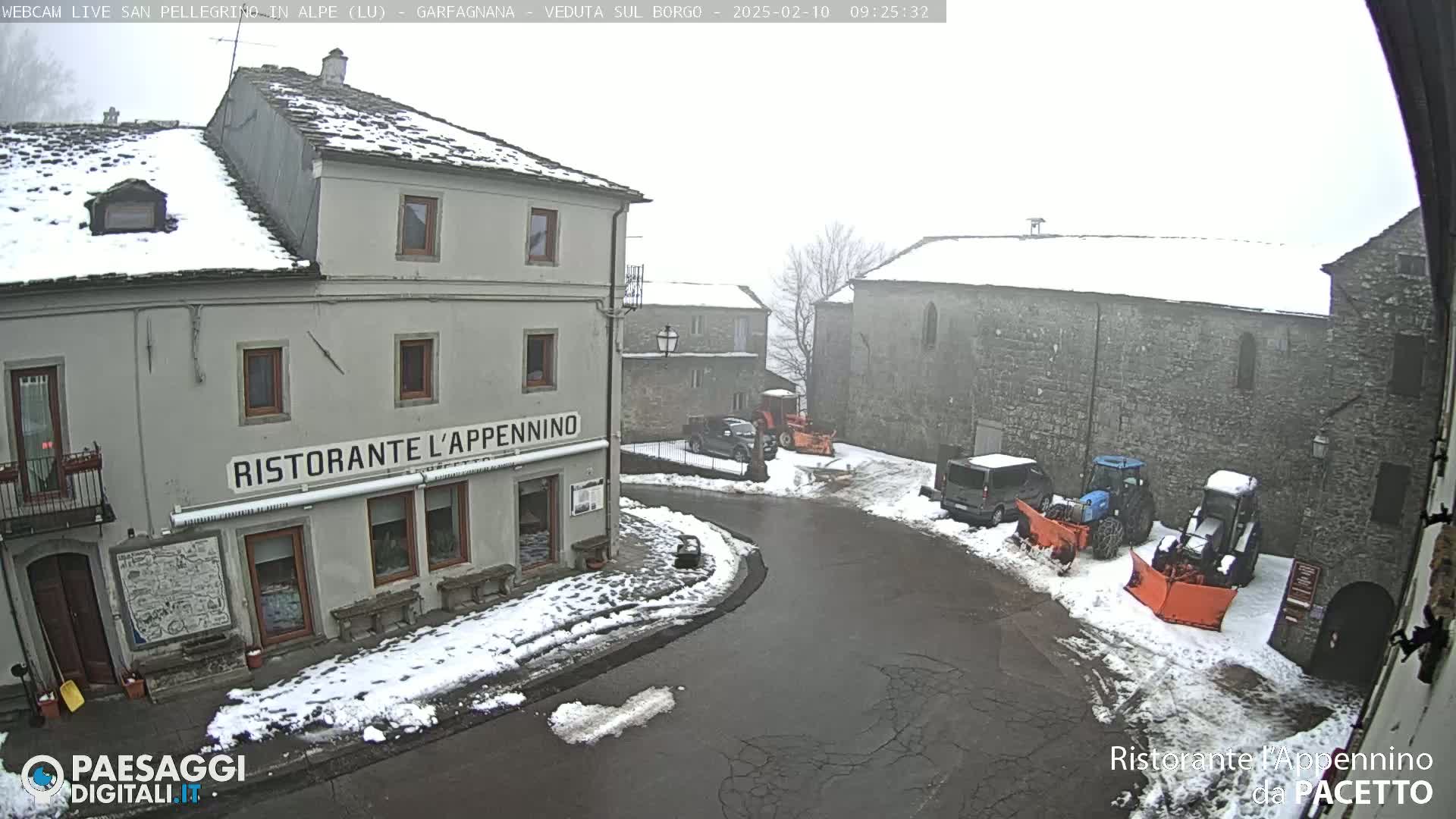 A snow-covered village square with several stone buildings, some vehicles, and snow plows.