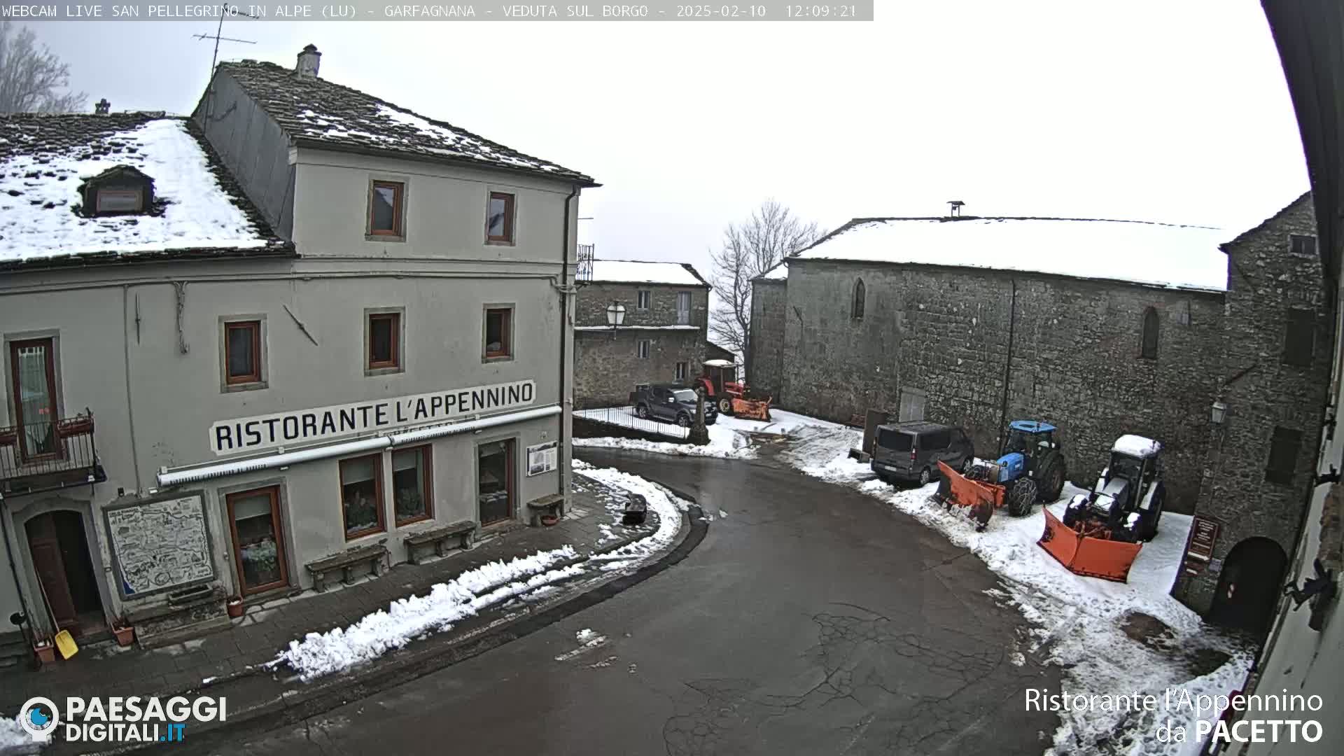 A snow-covered village square features several buildings, parked vehicles, and snow-clearing equipment under an overcast sky.