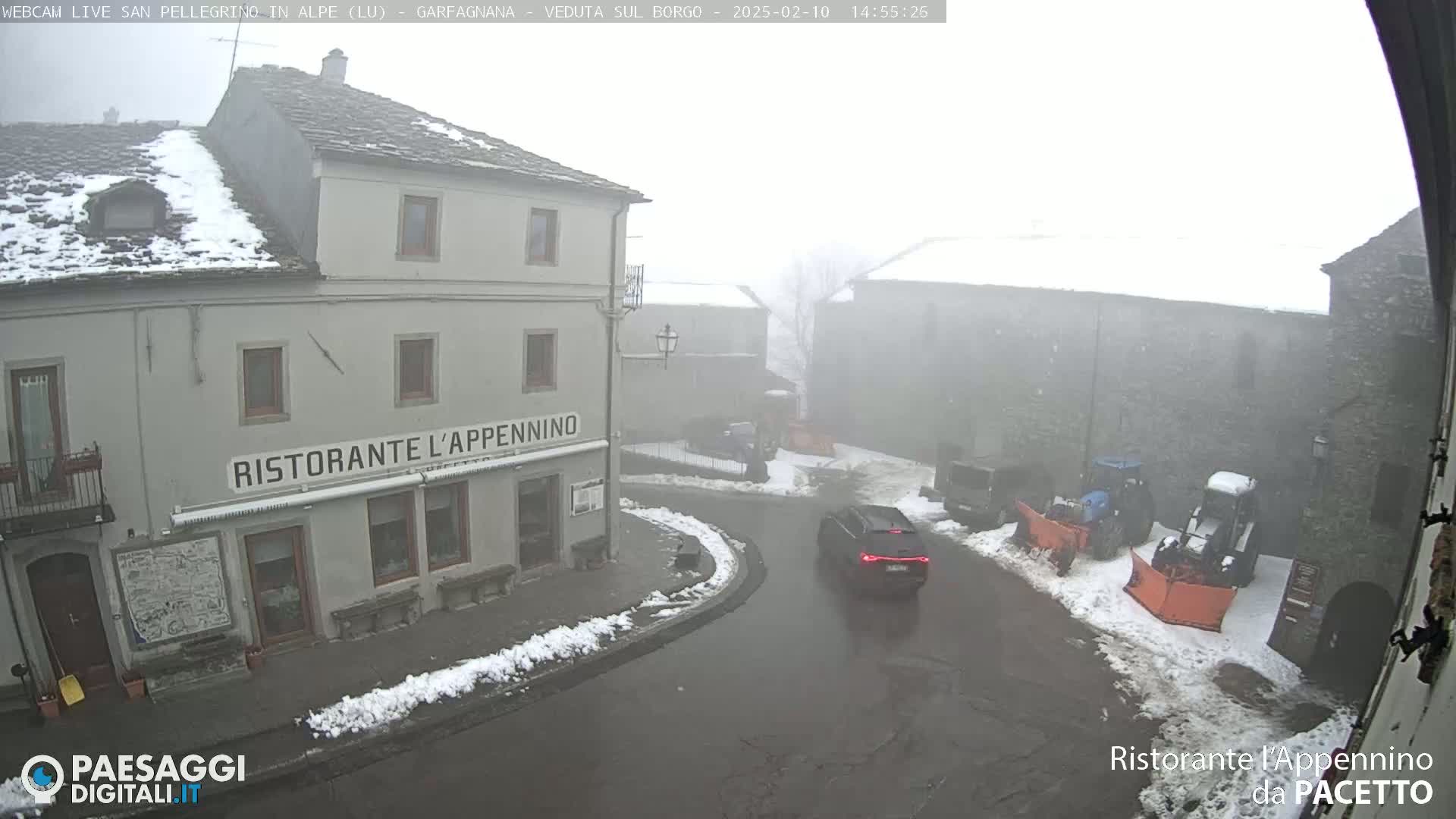 A foggy, snowy town square with a restaurant and several vehicles, including a snowplow.