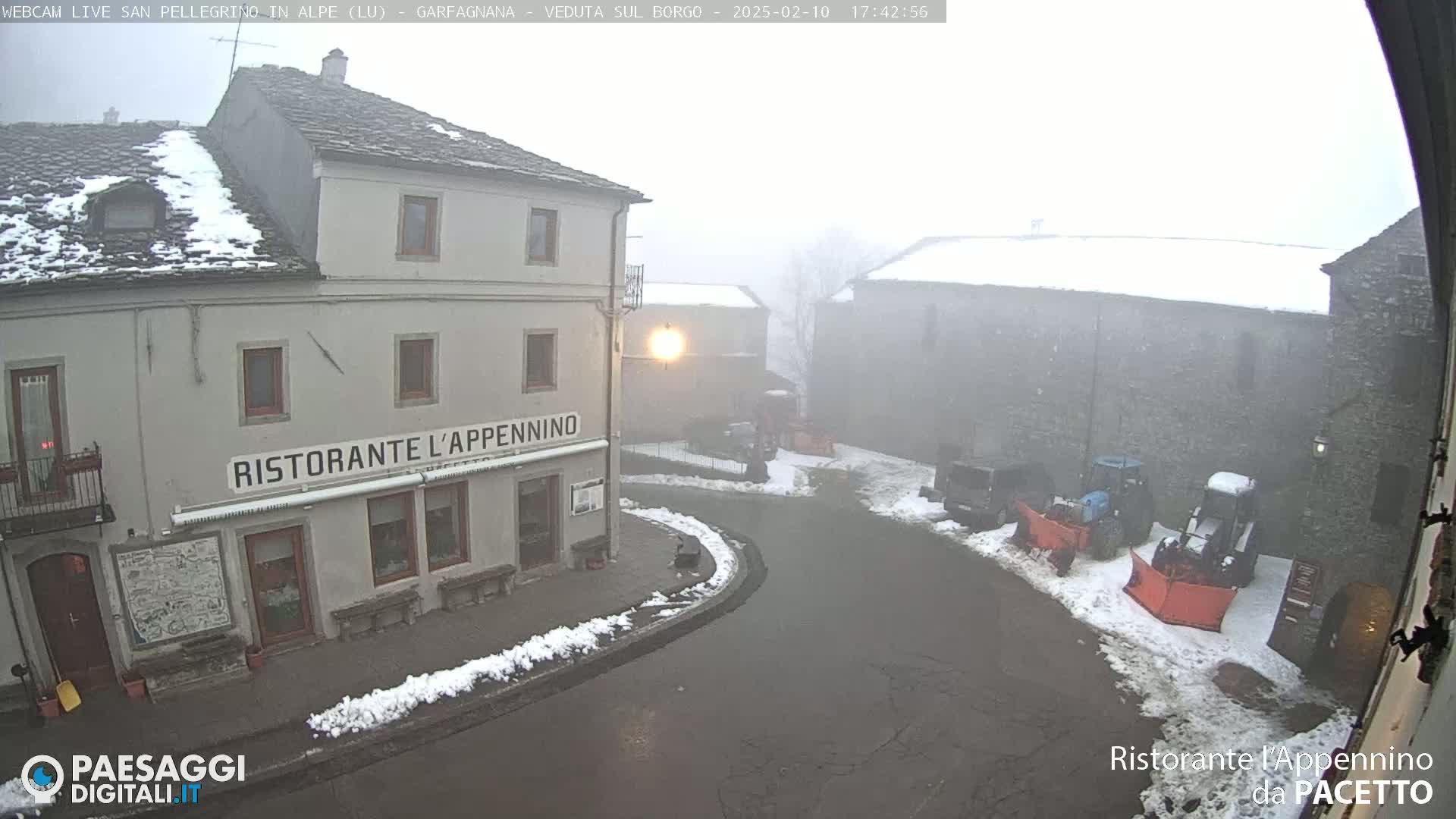 A snow-covered village square is viewed on an overcast day, with several stone buildings and snow-clearing equipment visible.