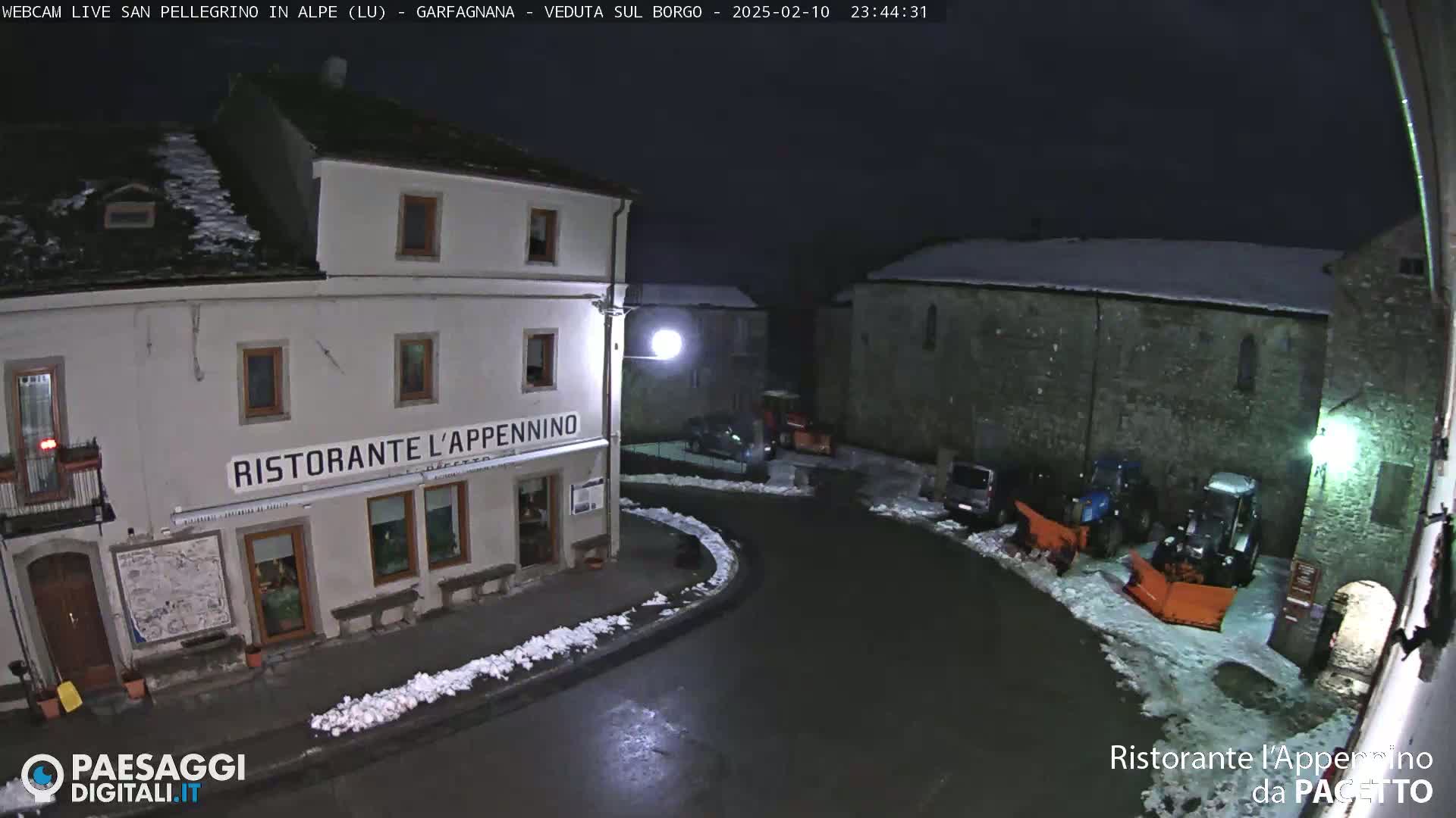 A snow-covered village square at night, illuminated by streetlights, features a restaurant, parked cars, and snow-removal equipment.
