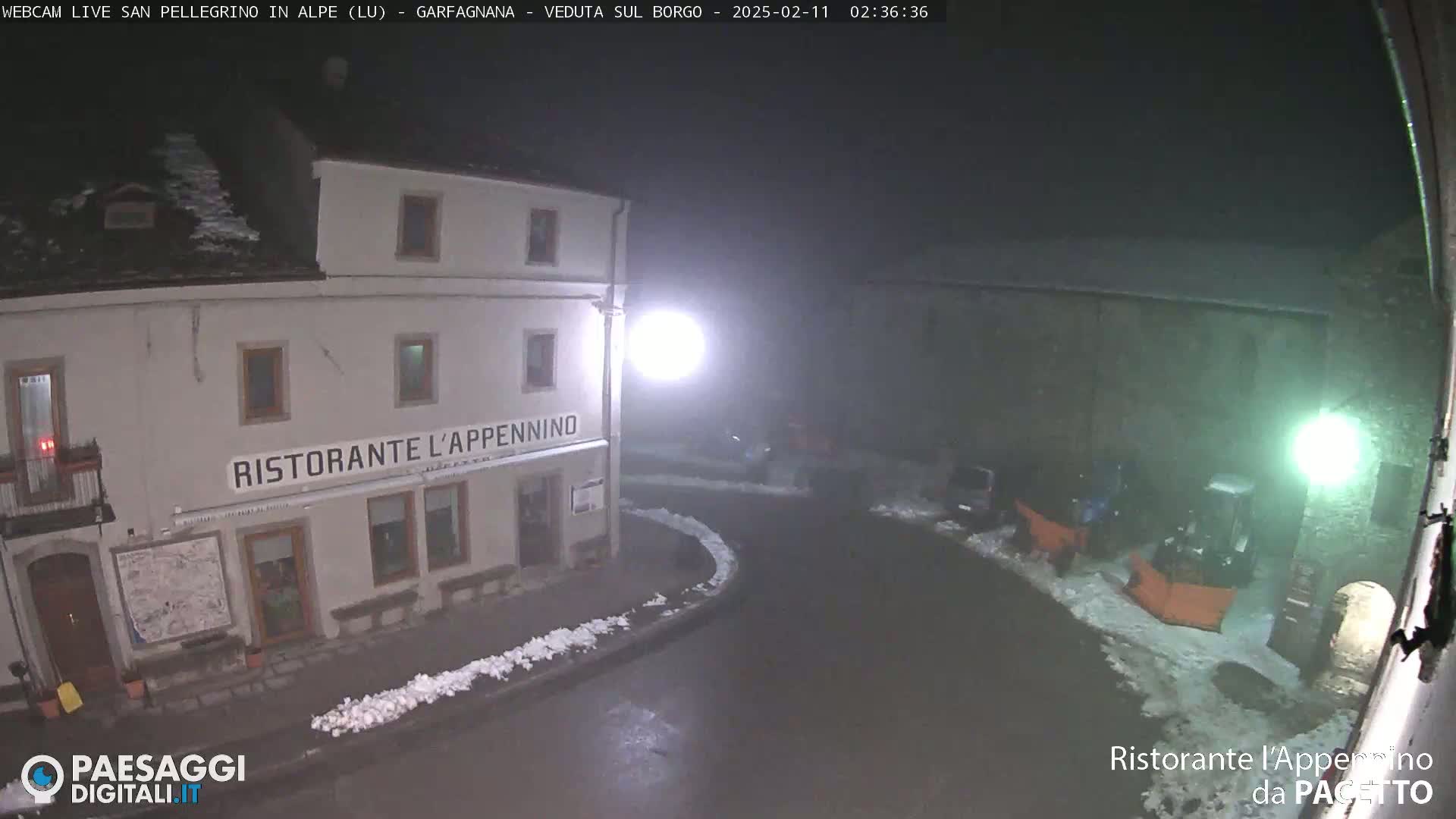 A nighttime view of a town square with snow on the ground, showing a restaurant and some parked vehicles, lit by street lamps under a dark, overcast sky.