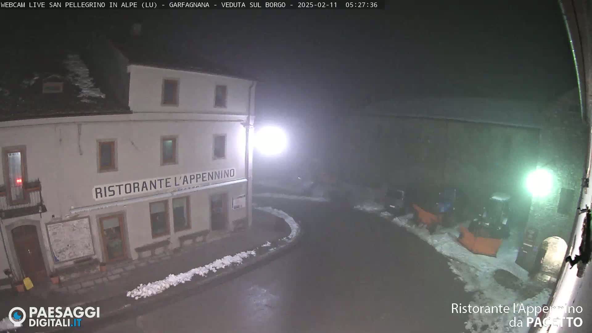 A nighttime view of a snow-covered town square with a restaurant and snowplows, under foggy conditions.