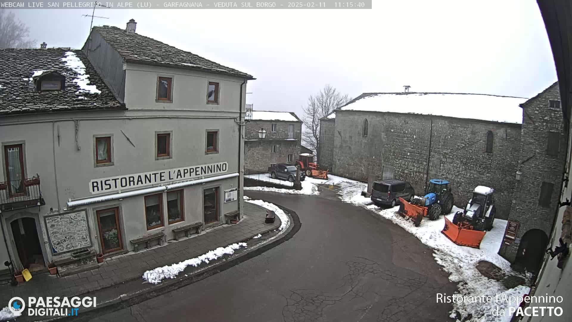 A snow-covered village square with several buildings, including a restaurant, and some parked vehicles, including tractors with snowplows.
