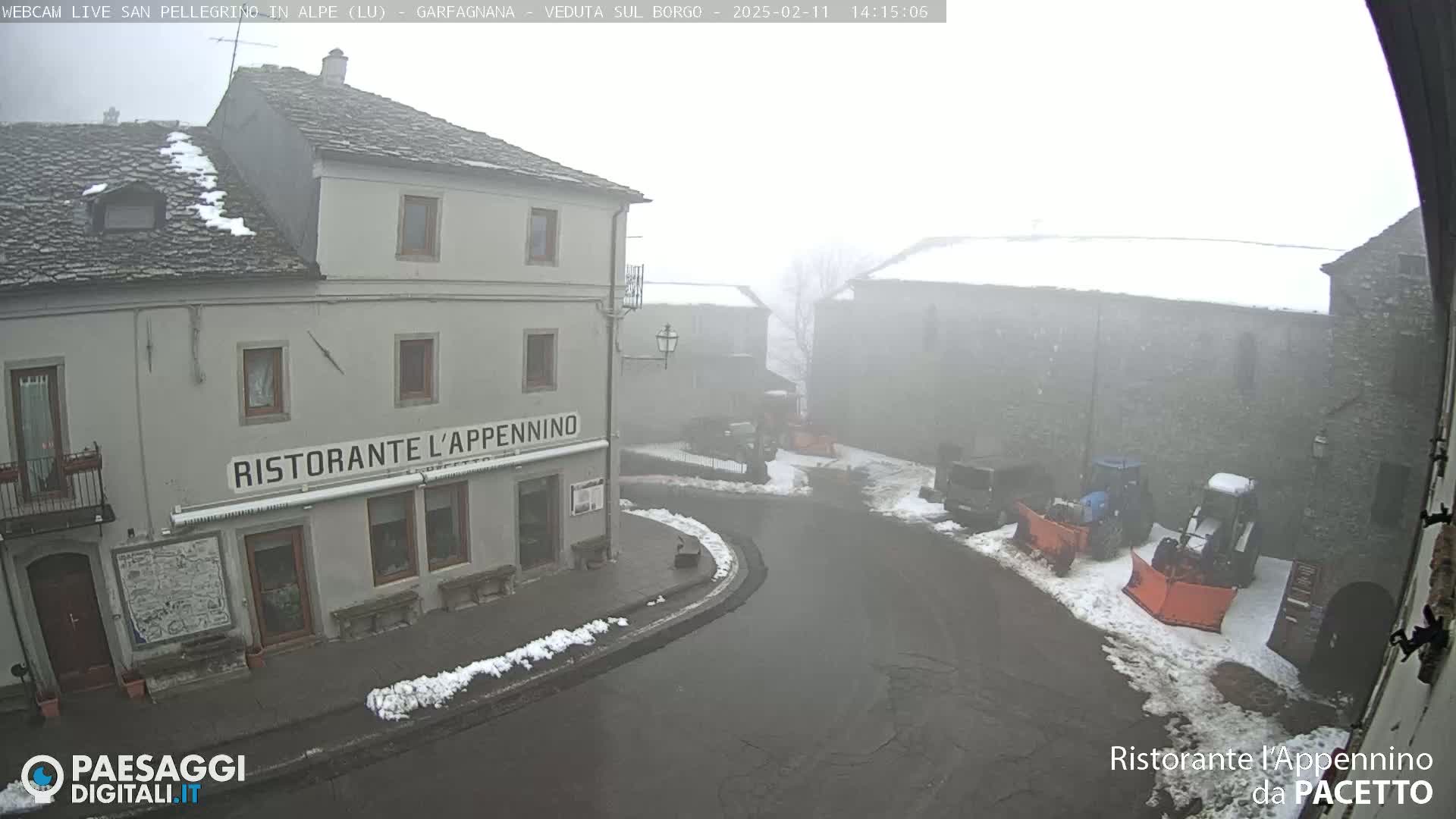 A foggy, snowy village square with a restaurant and several snow-covered vehicles.