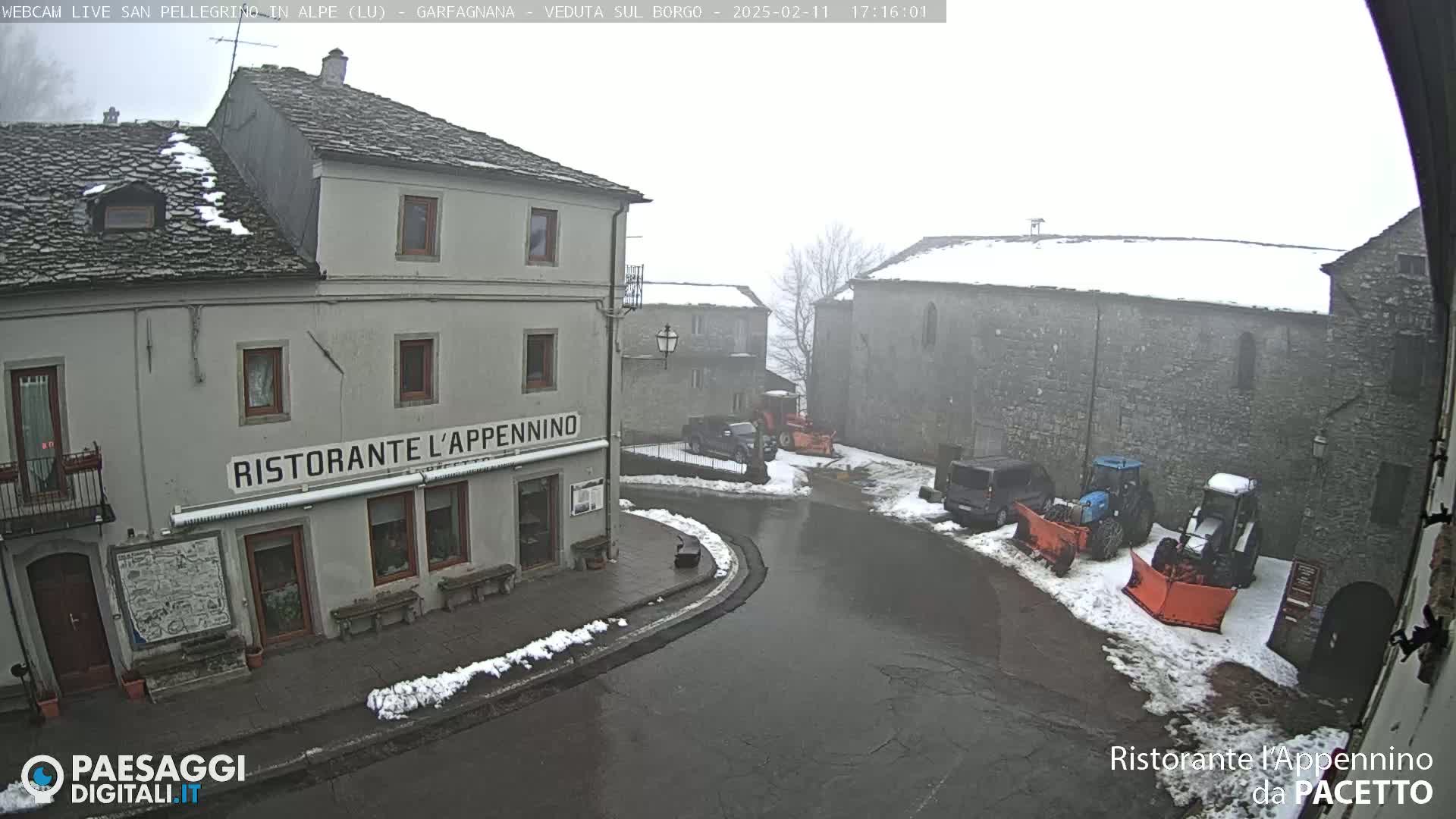 A snowy village square with several buildings, including a restaurant, and snow plows parked in the square under an overcast sky.