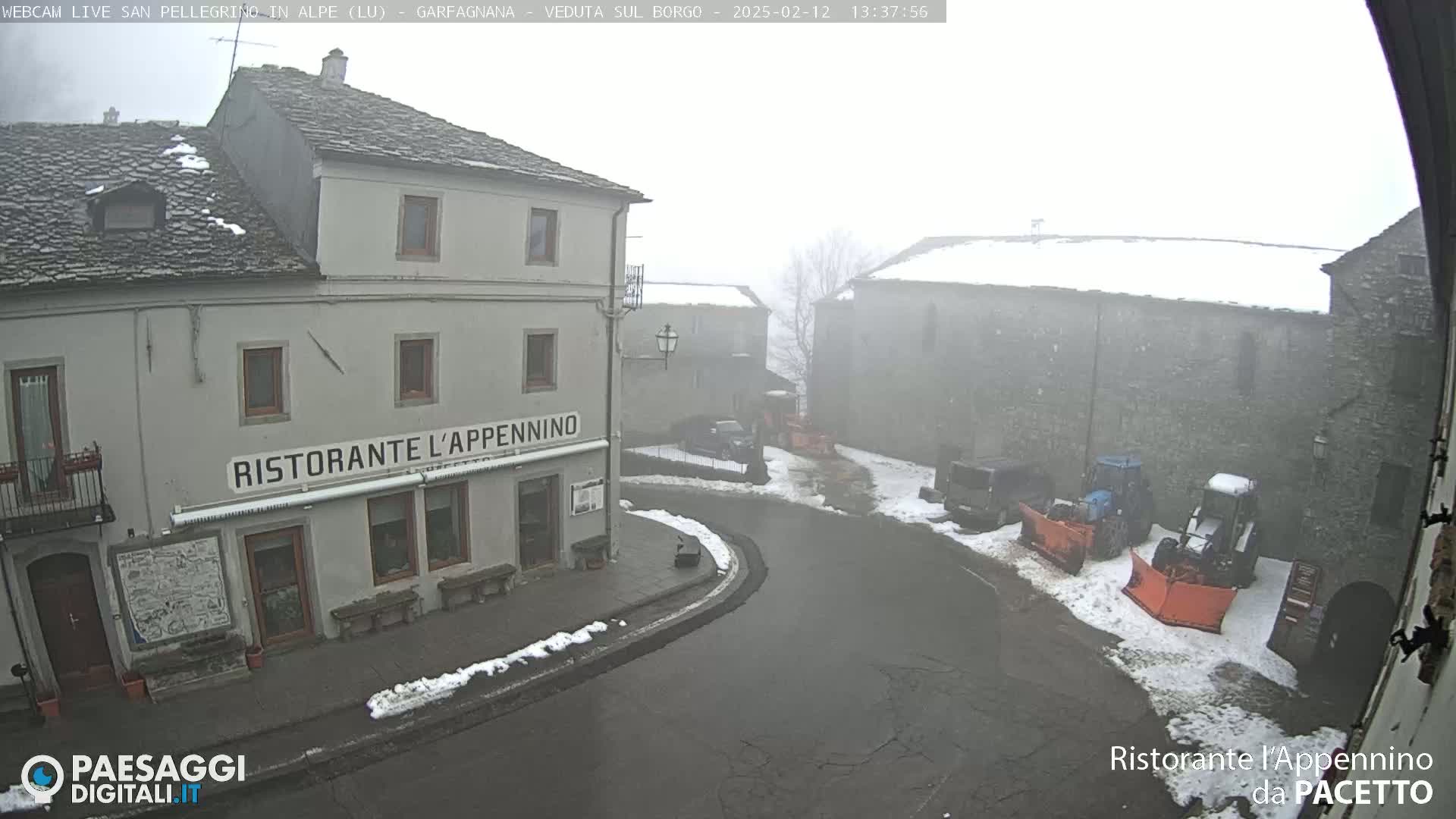 A foggy, snowy village square with stone buildings, some snow on the ground, and snow-clearing equipment.