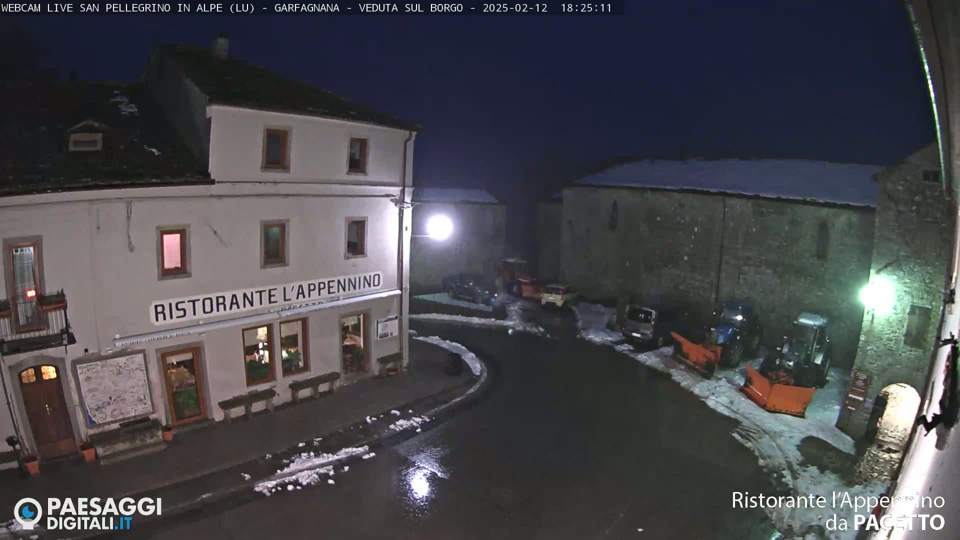 A nighttime view of a town square with snow on the ground, showing a restaurant and several parked vehicles, including snowplows.