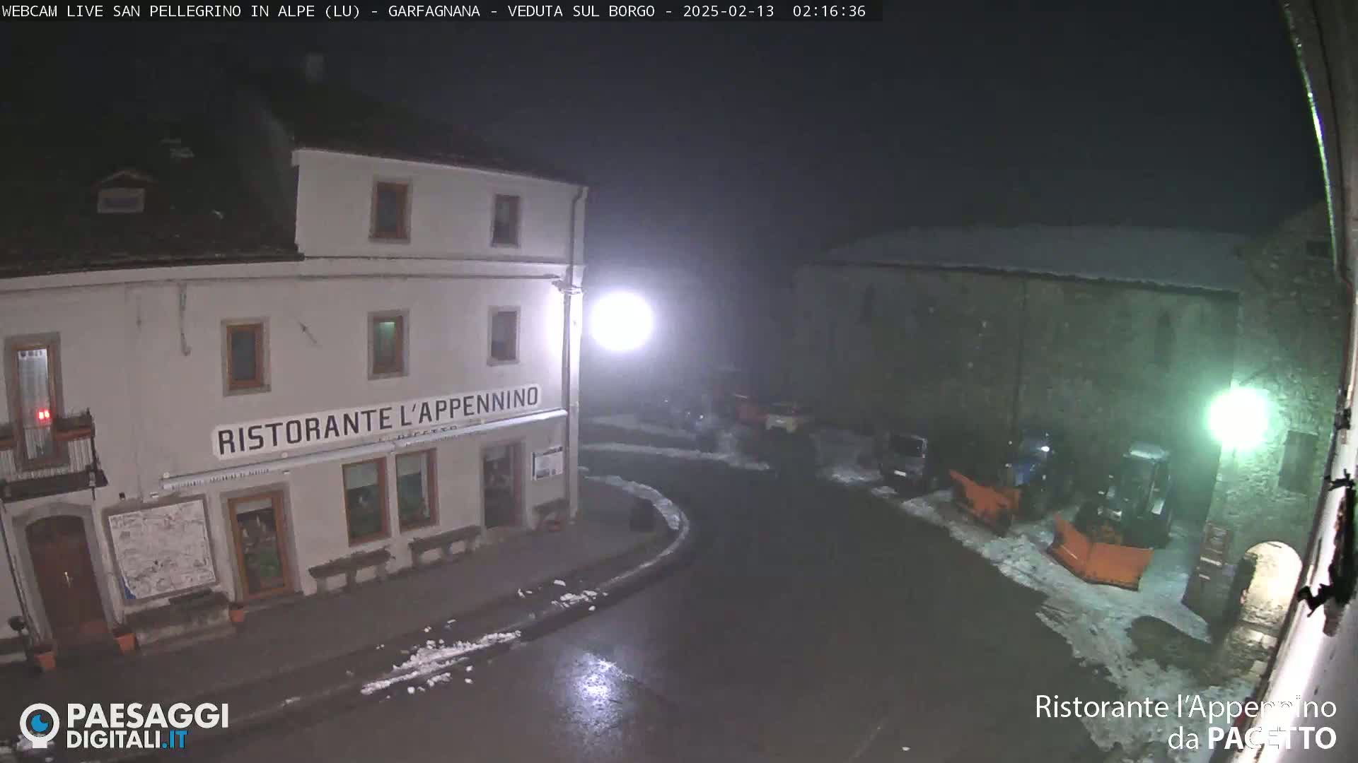 A nighttime view of a snow-dusted town square with a restaurant, some parked vehicles, and a snowplow.