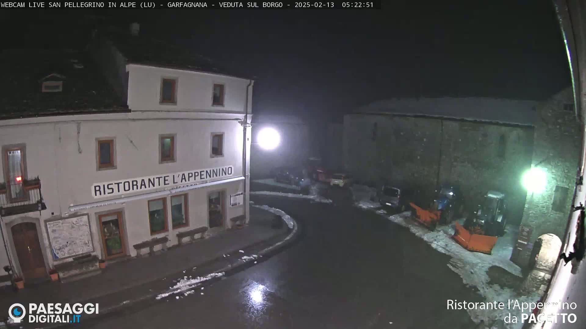 A nighttime view of a town square with snow on the ground, featuring a restaurant and snowplows parked nearby.
