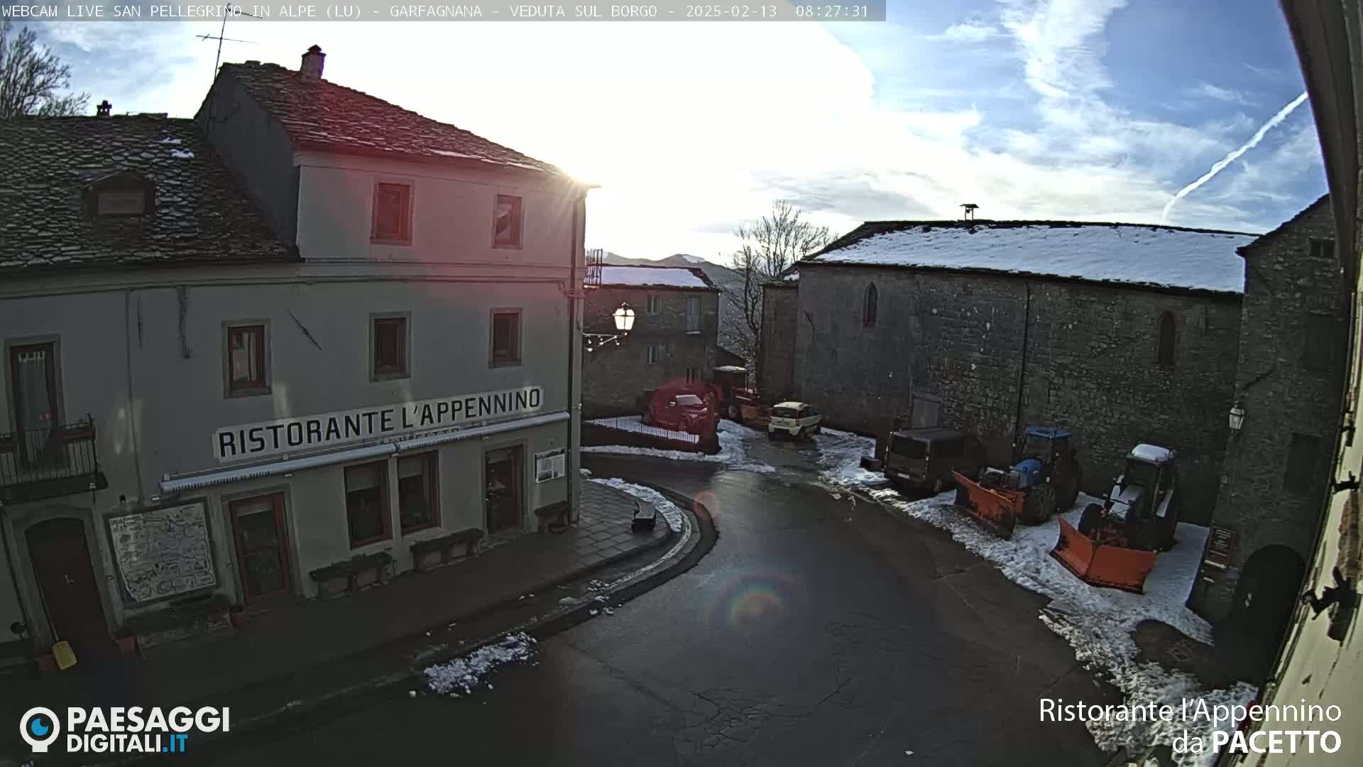 A sunny, partly cloudy day in a snow-dusted village square with several parked vehicles near a restaurant.