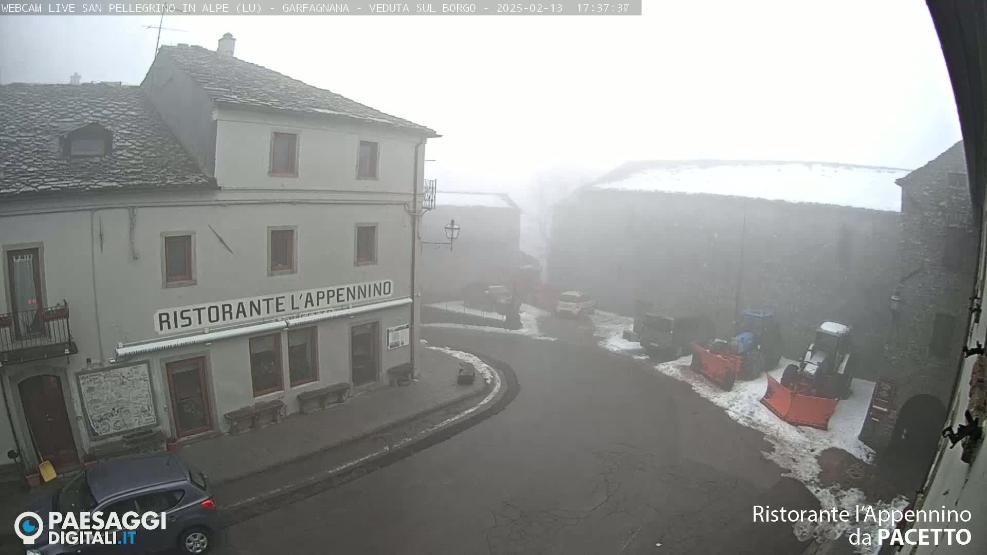 A foggy village scene shows a stone building with a restaurant, a street, and snow-covered ground with snowplows.