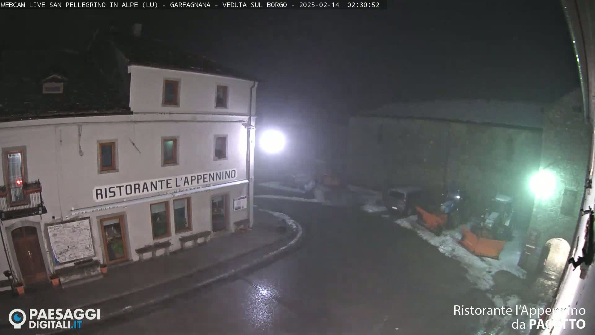 A nighttime view of a snow-covered town square with a restaurant, illuminated by streetlights, in snowy conditions.
