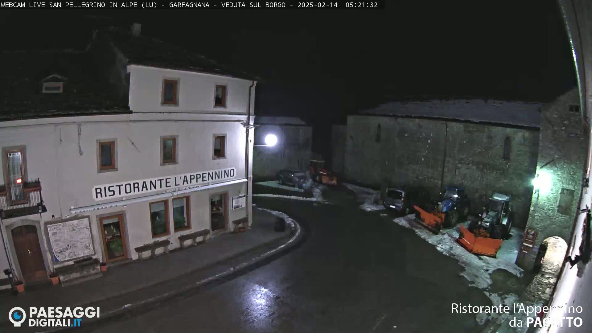 A nighttime view of a snow-dusted town square with parked vehicles and snow removal equipment near a restaurant.