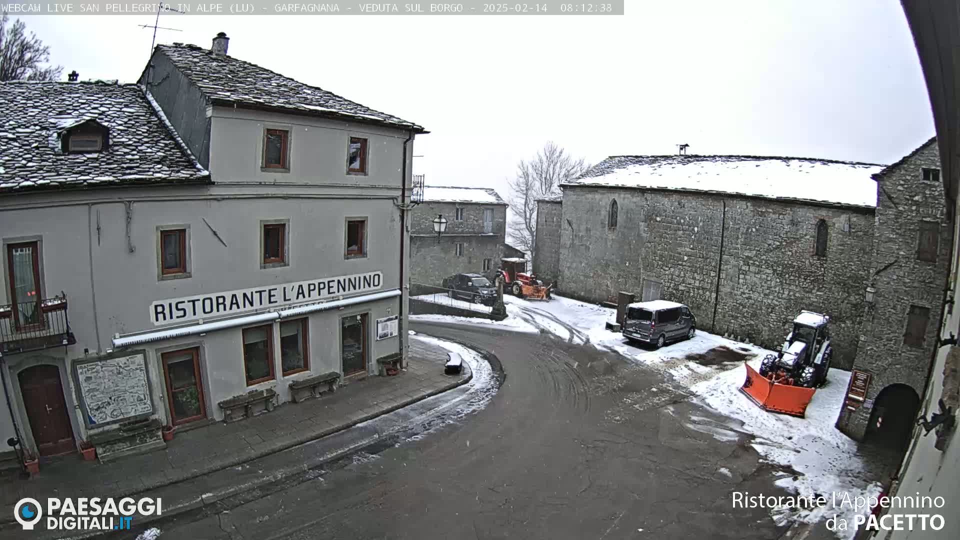 A snow-covered village square with stone buildings, a restaurant, several vehicles, and a snowplow.