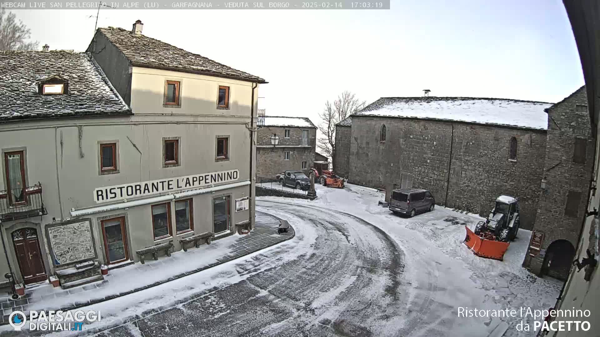 A snow-covered town square with several buildings, a few parked vehicles, and a snowplow.