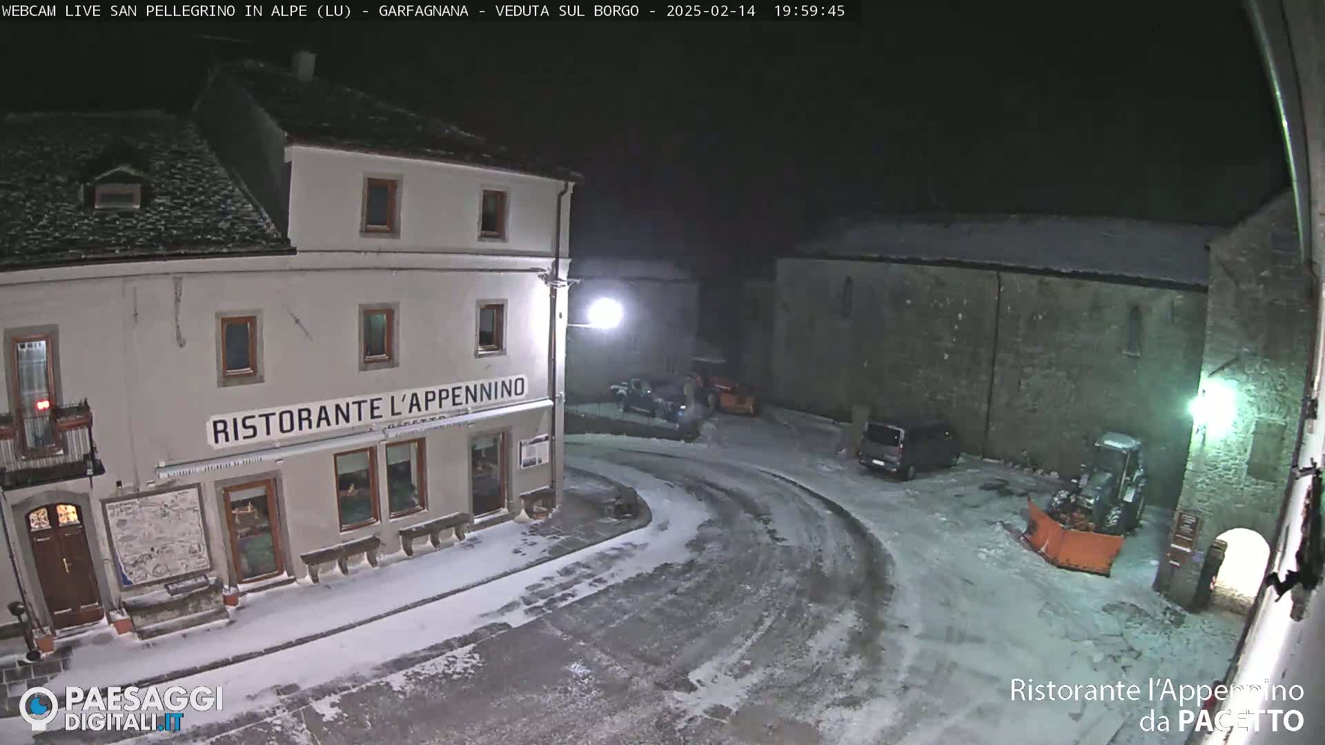 A snow-covered town square at night, featuring a restaurant and parked vehicles, with a snowplow visible.