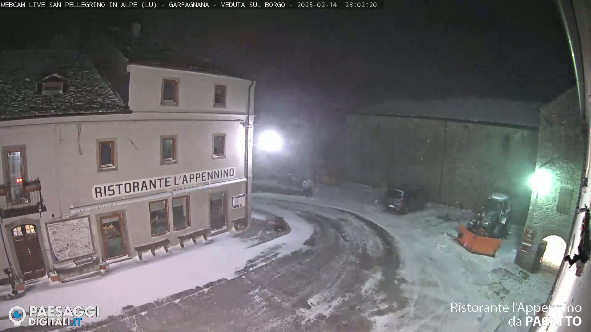 A snow-covered town square at night, with a restaurant and a snowplow visible, under light snowfall.