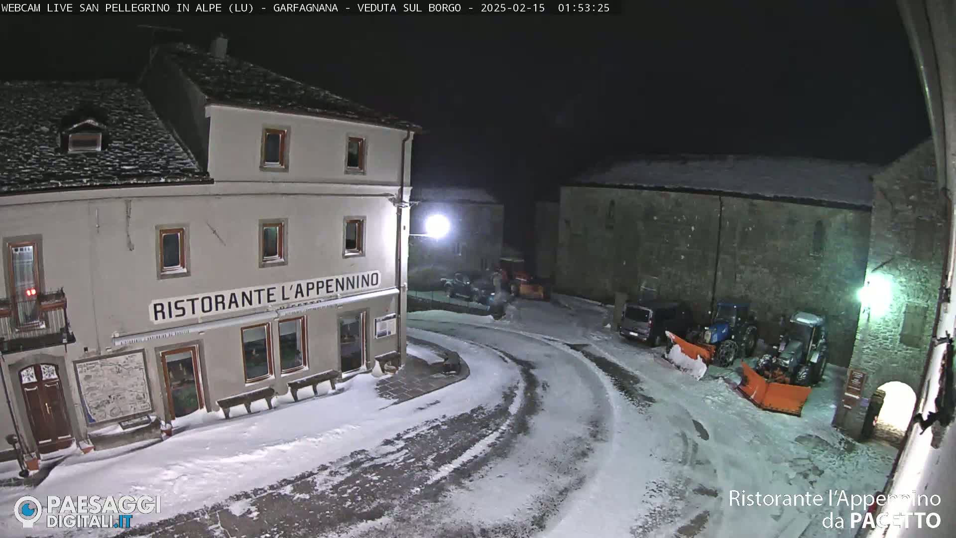 A snow-covered town square at night, with two tractors equipped with snowplows parked near a restaurant.
